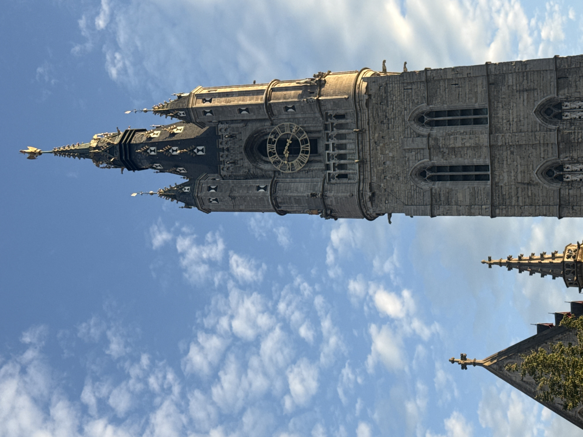 This is a low-angle, medium shot of the Belfry of Ghent, a medieval tower in Ghent, Belgium. The tower, made of grey stone, dominates the right side of the frame. It features a large clock face with Roman numerals and golden hands, surrounded by decorative architectural elements. The tower is topped with a golden dragon weather vane. To the left of the Belfry, the pointed roofs and spires of other gothic-style buildings are visible, including what appears to be a church with a cross on its roof. The sky is a clear blue, dotted with scattered white, fluffy clouds. The lighting suggests it is either early morning or late afternoon, with the sun casting a warm glow on the side of the tower. No people are visible in the frame.