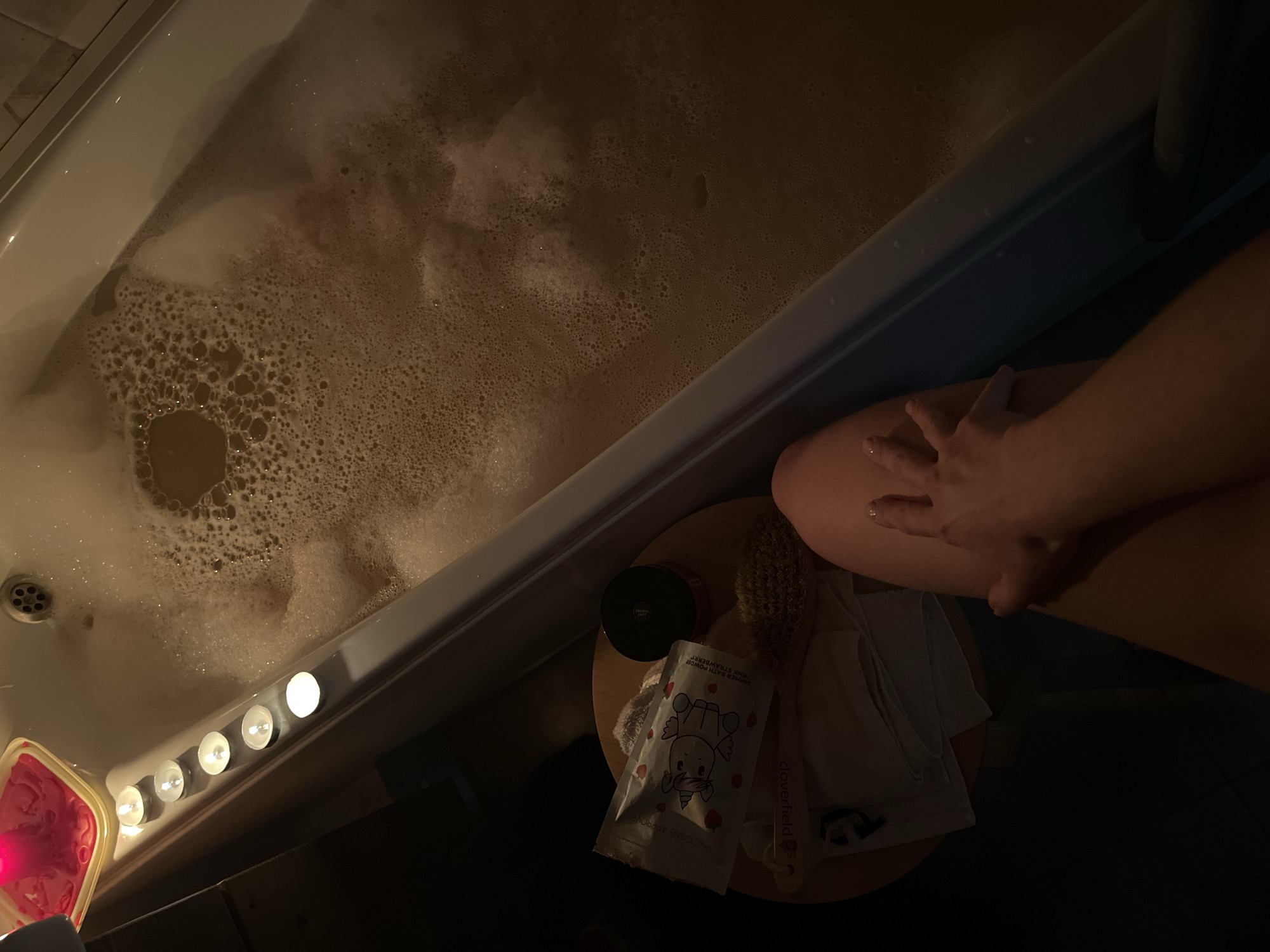 A dimly lit, high-angle shot shows a bathtub filled with bubbly, brown water. Several tea light candles are arranged along the edge of the tub, casting a warm glow. To the left of the tub, on a small wooden stool, are several bath-related items, including a packet with a unicorn design, a black jar, a body brush, and folded towels. A person's legs and hand are visible in the foreground, suggesting they are either entering or exiting the bath. The setting appears to be a bathroom, possibly in a residential dwelling in Kazan, Russia, given the implicit context of the request, although no specific Kazan landmarks or indicators are visible within the frame.