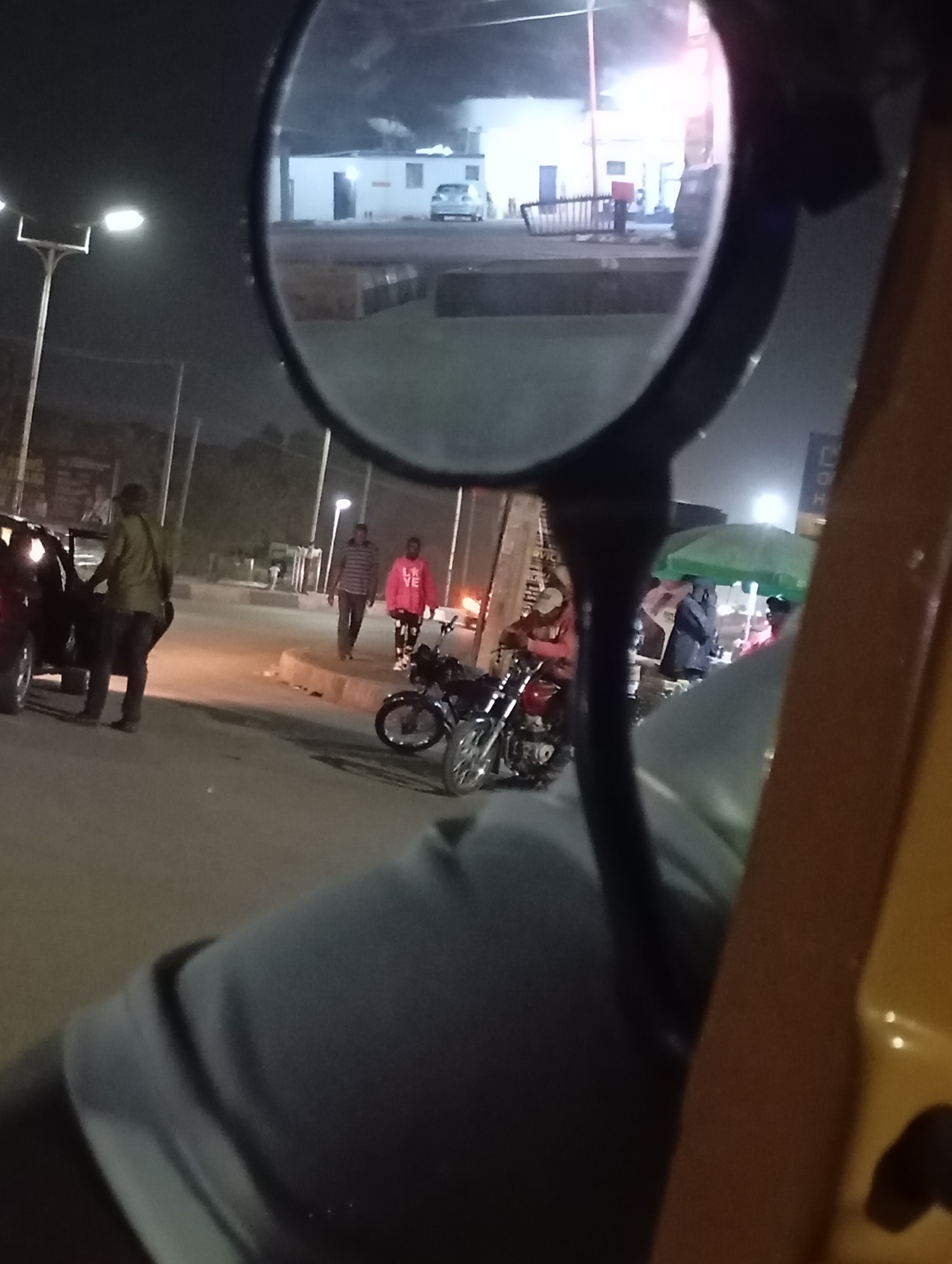 A nighttime scene in Jos, Nigeria, captured from the perspective of a passenger within a yellow vehicle, possibly a taxi or auto-rickshaw. The view is partially obscured by the vehicle's side mirror, which reflects a building, a light-colored car, and what appear to be industrial containers.
On the road, visible through the windshield and around the side mirror, several people are present. One man in a khaki jacket stands by an open car door. Two individuals, one in a striped shirt and the other in a pink sweatshirt, walk on the sidewalk. Motorcycles are parked along the roadside. A makeshift food stall with a green umbrella is in operation. Streetlights illuminate the scene.