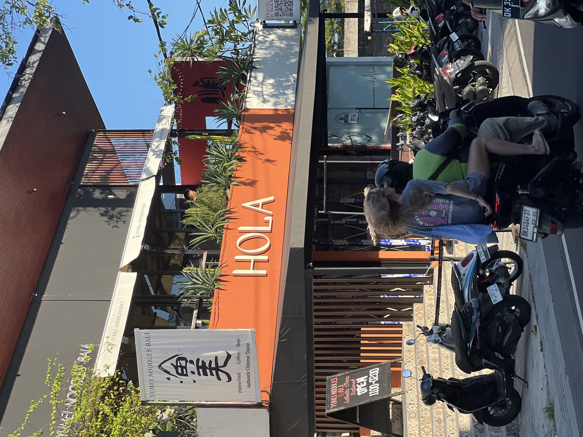 The image depicts a daytime scene in North Kuta, Indonesia, showcasing the exterior of "Yumei Noodles Bali" restaurant and "HOLA." The restaurant has an orange facade with the name "HOLA" written vertically and planted greenery. A vertical white banner with the text "Yumei Noodles Bali" is displayed next to the storefront, along with a small sign indicating opening hours from 11:00 to 02:00.
Several motorcycles are parked along the street in front of the establishment. Two individuals are seated on one of the motorcycles. One wears a blue shirt and a blue helmet, while the other wears a green shirt.