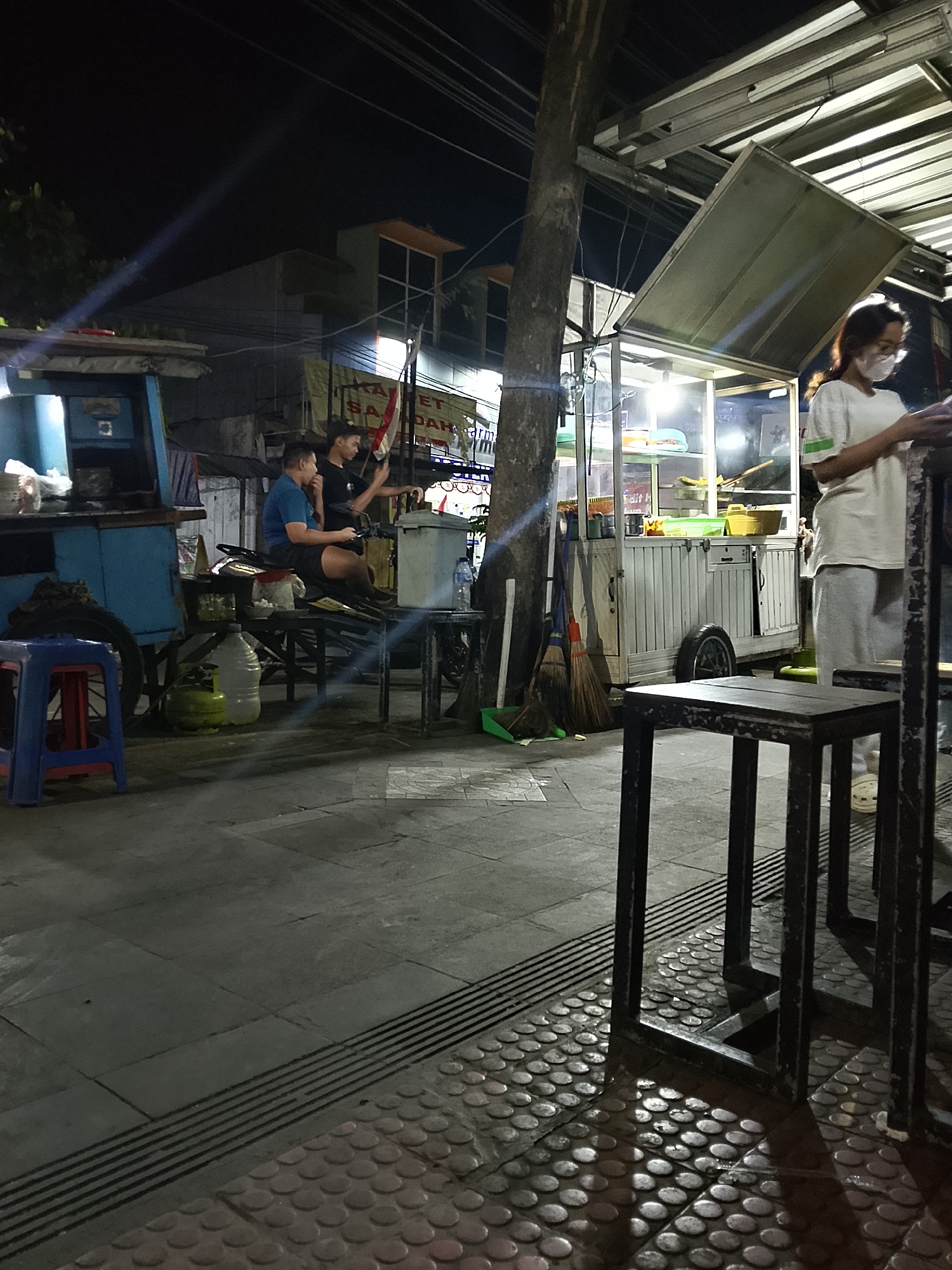 The image depicts a night scene at a street food stall, likely in Balun, Indonesia. A woman wearing a face mask and a light-colored top and pants stands behind a food cart, illuminated by overhead lights. Two men are seated on a motorcycle in front of another food cart. The man on the left wears a blue shirt and black shorts, while the man on the right wears a black t-shirt and black shorts. A third person, partially visible, is also near the men. Several objects are present, including brooms, a green gas tank, water bottles, and various items on the food carts. The ground is paved with textured tiles. The lighting suggests it is nighttime, with artificial lights from the stalls casting shadows.