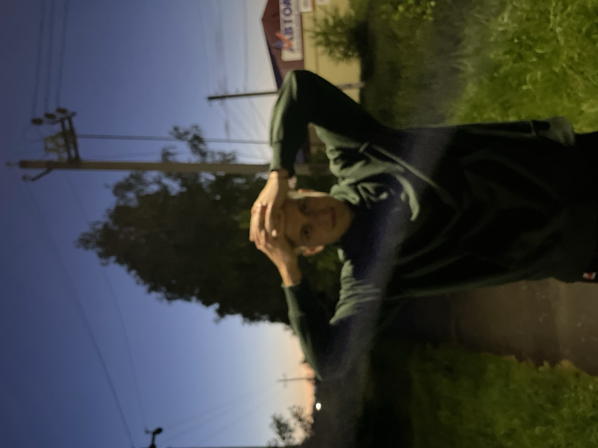 The image, taken in Красные Баки, Russia, captures a young individual posing outdoors during twilight or early evening. The photograph is oriented with a 90-degree clockwise rotation.
The central subject is a young person, likely male, wearing a dark green or black hooded sweatshirt. They are positioned facing the camera, with both hands raised to frame their head and face. Their expression appears neutral or subtly smiling.
The setting is an outdoor area, possibly near a street or sidewalk. The background features a fading blue and lighter orange sky, indicating dusk or dawn. Several utility poles with electrical wires are visible, suggesting an urban or suburban landscape. Dark trees and foliage are scattered across the scene, with distinct green grass or bushes lining the ground on the right. In the upper background, part of a building is visible, featuring a red and white sign. The visible text on the sign, in Cyrillic, reads "АВТОМ," suggesting it might be part of "Автомагазин" (Car Store) or a similar establishment.
The activity depicted is the person posing for the camera. The overall atmosphere is calm and still, with no dynamic action taking place. The lighting is low, characteristic of the transition period between day and night. The weather appears clear, with no clouds visible.