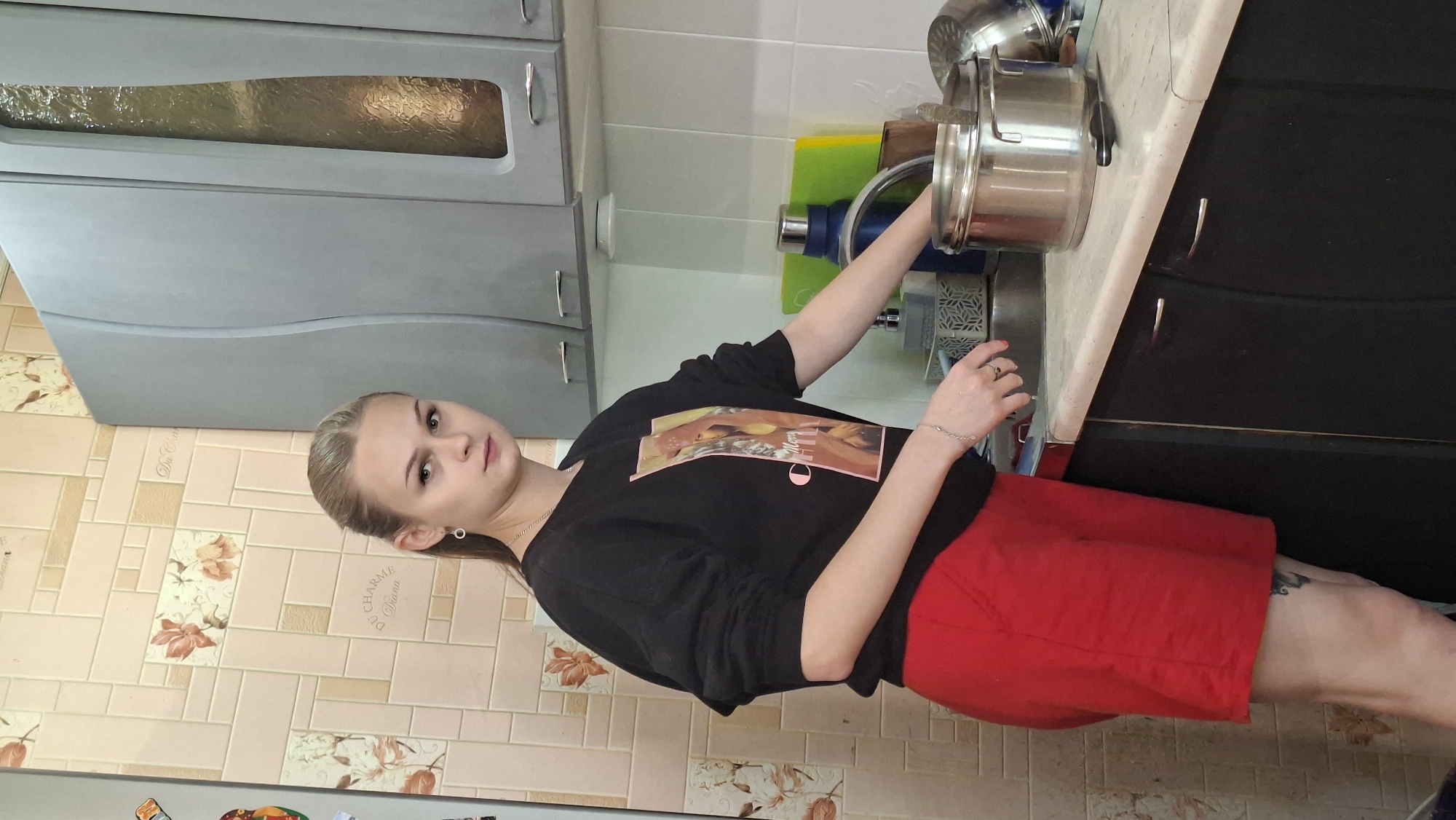 A young woman with blonde hair pulled back in a ponytail is standing in a kitchen. She is wearing a black sweatshirt with a graphic on the front and red shorts. She is leaning over the sink, with her right arm extended, appearing to be washing dishes. There are pots and pans on the counter next to the sink. The kitchen has beige and pink floral patterned tiles on the backsplash and gray cabinets. The scene is likely indoors in a residential setting. The location is specified as Myadzyel, Belarus.