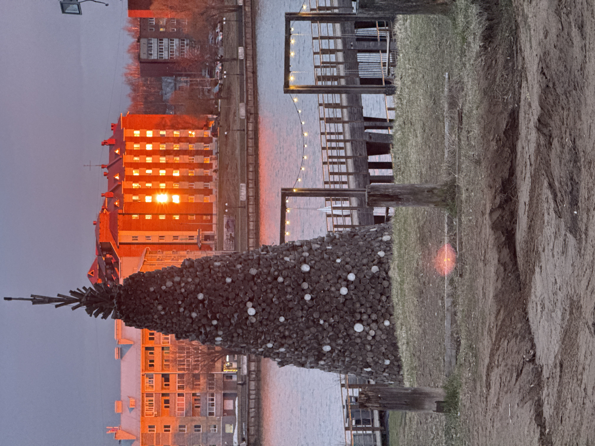 An evening scene in Votkinsk, Russia, captures a waterfront view at dusk or dawn. In the midground, a large, conical sculpture made of stacked logs or wooden discs, resembling a Christmas tree, stands on a grassy bank. This unique "tree" is adorned with scattered white, spherical decorations. To its right, the ground transitions from grass to a rough, muddy embankment.
Across a calm body of water, possibly the Votkinsk Reservoir, the opposite bank features urban development. Several multi-story buildings are visible, with one prominent structure on the left bathed in a vibrant, warm orange light, suggesting either sunrise or sunset. Numerous windows in this building glow brightly with the same orange hue, reflecting the intense light. Further along the bank, docks or jetties extend into the water, strung with decorative lights that cast subtle reflections. The sky above is a muted grey-blue, providing a cool contrast to the warm light on the buildings and water. A street light with a floodlight is visible in the upper left. The overall atmosphere is tranquil, highlighting the interplay of natural and urban elements under the striking glow of the setting or rising sun. There are no people visible, and no discernible text.