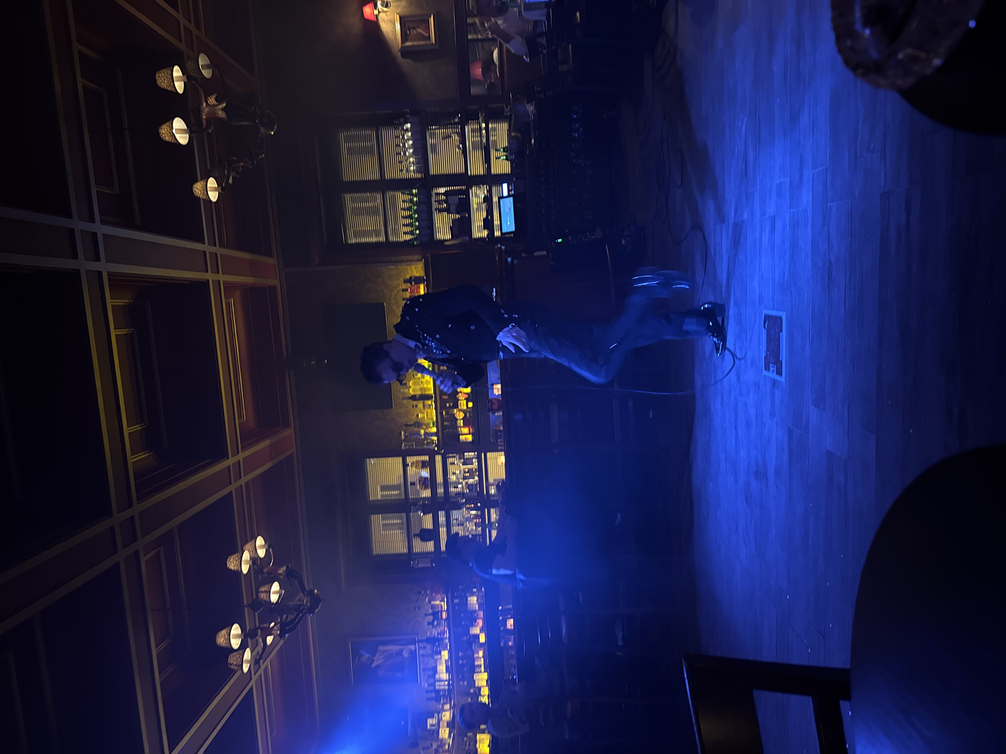 Here is a description of the image:
The image shows a man standing on a wooden floor in what appears to be a bar or club. The scene is illuminated by blue lighting, creating a dimly lit and atmospheric environment. In the background, there is a bar with shelves stocked with bottles, suggesting a place where drinks are served. There are also windows with blinds in the background. The man is dressed in a dark, possibly sequined jacket and is holding a microphone. The presence of the microphone indicates the man might be performing. Chandeliers are visible on the left side of the image, adding to the decorative atmosphere of the location. Several tables are visible, and a chair is in the lower right corner.