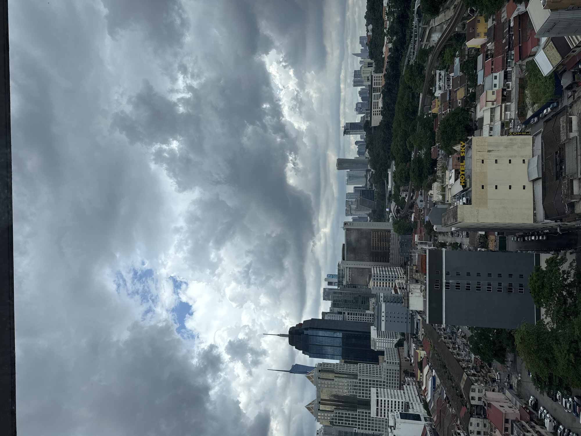 The image captures a panoramic view of Kuala Lumpur, Malaysia, under a dramatic sky filled with dark, heavy clouds, with a small patch of blue sky visible in the upper center. The cityscape is dominated by a dense collection of skyscrapers of varying heights and architectural styles. In the foreground, a dark, imposing skyscraper with a pointed roof and a dark facade stands out. To its left, a cluster of lighter-colored buildings features reflective glass facades and angular tops. Further into the distance, more buildings form a continuous urban horizon, some with distinctive shapes, such as the tall, slender skyscraper with a blue spire in the mid-ground.
Below the skyscrapers, a more suburban or older section of the city is visible, characterized by lower-rise buildings with red-tiled roofs, some appearing weathered. Streets with parked cars are interspersed among these residential or commercial structures. A prominent building in the mid-ground features a sign that reads "HOTEL SKY" in yellow capital letters. Greenery, in the form of trees and parks, is scattered throughout the city, providing a contrast to the urban development. A railway track and a bridge are also discernible in the middle ground, indicating transportation infrastructure. The overall impression is a bustling metropolis under an atmospheric, possibly pre-rainy, sky. There are no discernible people or specific actions taking place in the scene.