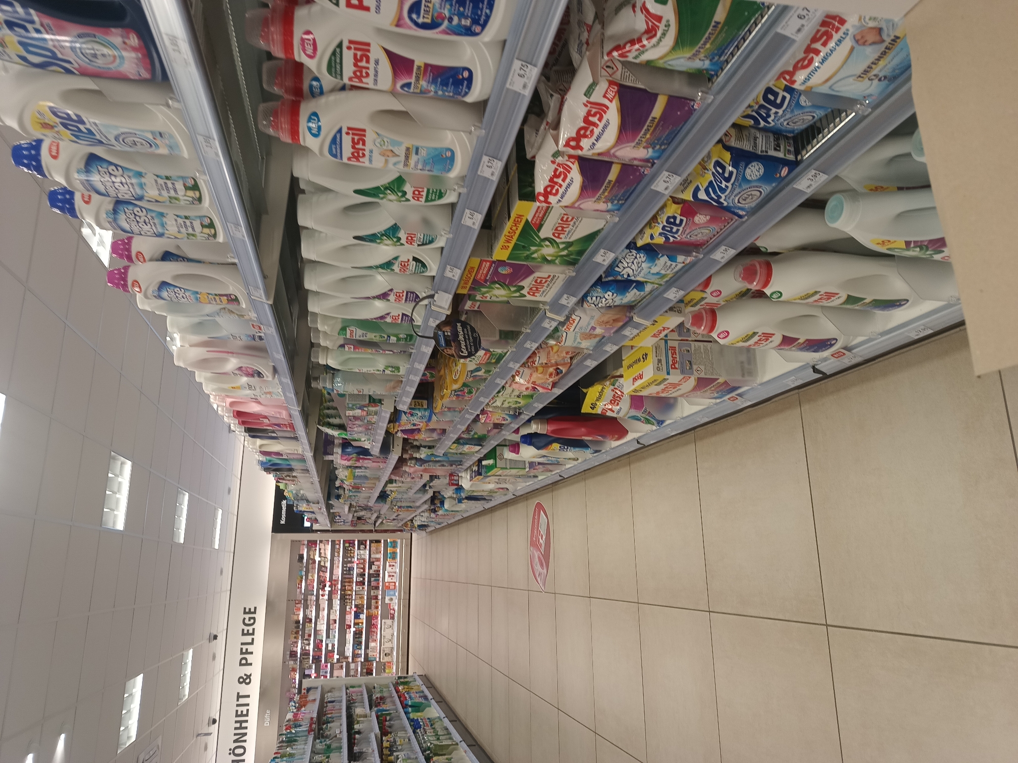 This is a photograph of a retail store aisle, likely a supermarket or drugstore, with a focus on laundry detergents and cleaning supplies. The shelves are heavily stocked with various brands and sizes of detergent bottles, boxes, and pods. Prominent brands visible include Persil and Ariel, with German text indicating product features like "Tiefenrein" (deep clean) and "Megaperls" (mega pearls). There are also smaller bottles and packets of other cleaning products.
The image is taken from a low angle, looking up along the aisle. The lighting appears to be typical indoor fluorescent lighting. The floor is tiled with large, light-colored square tiles. On the right side of the aisle, a red sign with a white pictogram is visible on the floor, possibly indicating a sale or promotion.
In the background, beyond the cleaning supplies, there are displays of cosmetics and fragrances, as indicated by the text "Kosmetik" and "Düfte" on a shelf header. On the left side of the image, a large sign reads "HÖNHEIT & PFLEGE" (Beauty & Care), further confirming the nature of the retail establishment. The overall impression is of a well-stocked retail environment, emphasizing household cleaning and personal care products. No people are visible in the frame. The date and time are not discernible from the image.