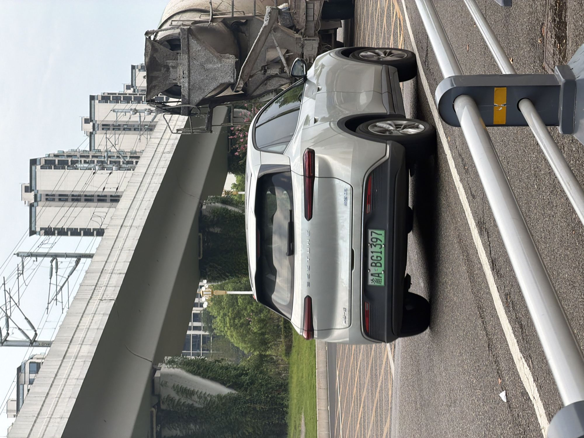 In the city of Jiangjin, China, a light-colored, four-door electric SUV, identified by its license plate "A BG1397," is captured in motion on an asphalt road. The vehicle's rear is prominently featured, showcasing its taillights and the "BESTUNE" branding. To the right of the SUV, a large concrete mixer truck is partially visible, suggesting a construction or industrial context. The background reveals a multi-story overpass supported by concrete pillars and a dense arrangement of tall, modern residential buildings, indicative of urban development. Overhead power lines are strung along the overpass, and lush green vegetation, including ivy, grows on the sides of the structure, indicating a well-maintained urban environment with ongoing construction or infrastructure projects. The sky is overcast, contributing to the ambient light of the scene.