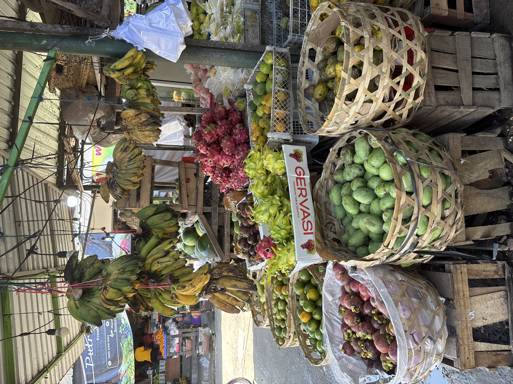 This image depicts a fruit stall in Ungasan, Indonesia. Bunches of green and yellow bananas hang from the stall's roof. Below, baskets and crates contain an assortment of fruits, including mangoes, dragon fruit (pink and yellow varieties), oranges, and mangosteens in plastic bags. A sign for "Gervasi Premium Table Grapes" is visible amidst the fruit displays. The stall is constructed of wood and metal, with a tarp on one side. Sunlight shines on the scene.