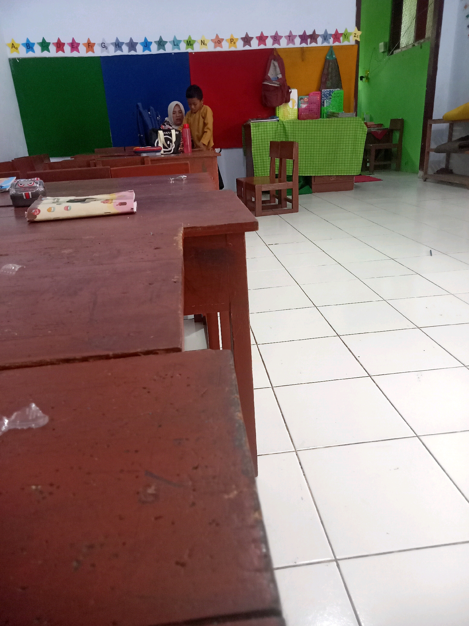 This media file depicts a classroom setting, likely in Pule, Indonesia, as suggested by the location context. The scene is filled with wooden desks, some in the foreground showing signs of wear and tear. In the background, a woman wearing a hijab and a young boy in a yellow shirt are visible behind a desk. They appear to be engaged in an activity, possibly related to education, as educational charts and colorful star-shaped decorations with letters of the alphabet are displayed on the wall behind them.
The room has a vibrant feel, with green, blue, and red panels on the wall, contributing to a lively atmosphere. There are also various objects on the desks, including a pencil case with a cartoon character, a red bottle, and other school supplies. A green tablecloth covers a table in the background, adorned with colorful storage bins and books. The floor is tiled with white squares, reflecting the ambient light, which suggests it might be daytime. The overall impression is that of a functional and engaging learning environment for young children.