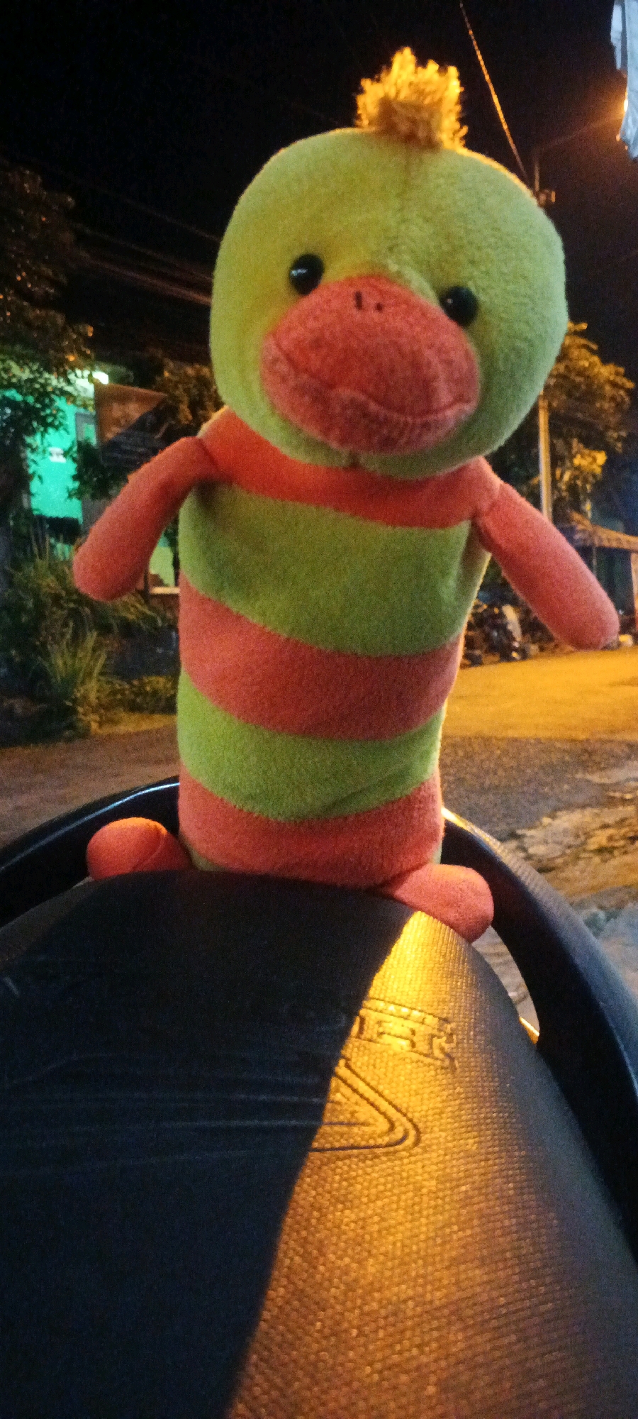 A stuffed toy duck with a green head and orange and green striped body stands on the black seat of a motorcycle. The duck's head is round with small black eyes and a large orange beak. It has tufts of yellow fluff on top of its head. Its arms are orange and rest by its sides. The duck's feet are also orange and appear to be tucked underneath it, resting on the seat. The motorcycle seat has a textured surface with a faint logo embossed on it, illuminated by an orange light source from the right. The background is dark, indicating it is nighttime, with some blurry foliage and what appear to be streetlights or building lights visible in the distance. The scene is likely outdoors, possibly on a street in Kesiman, Indonesia.
