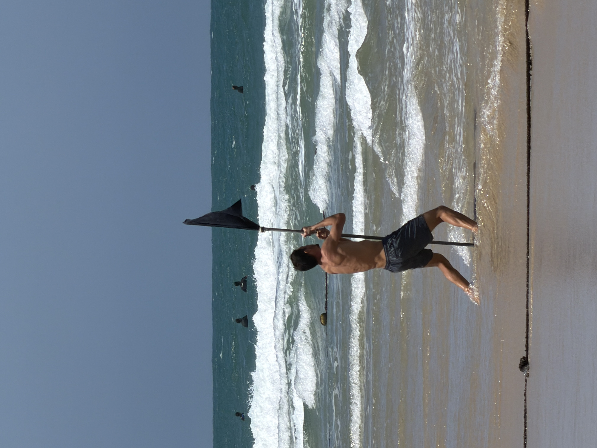 The image captures a man on a beach in what appears to be Tel Aviv, Israel. He's tanned, shirtless, and wearing dark shorts. He's waist-deep in the water and holding a tall, dark-colored flag. The beach is sandy with foamy waves washing ashore. In the background, the sea is a gradient of blues and greens, and there are several people swimming in the sea. The sky is light blue. The image appears to be taken during daylight.