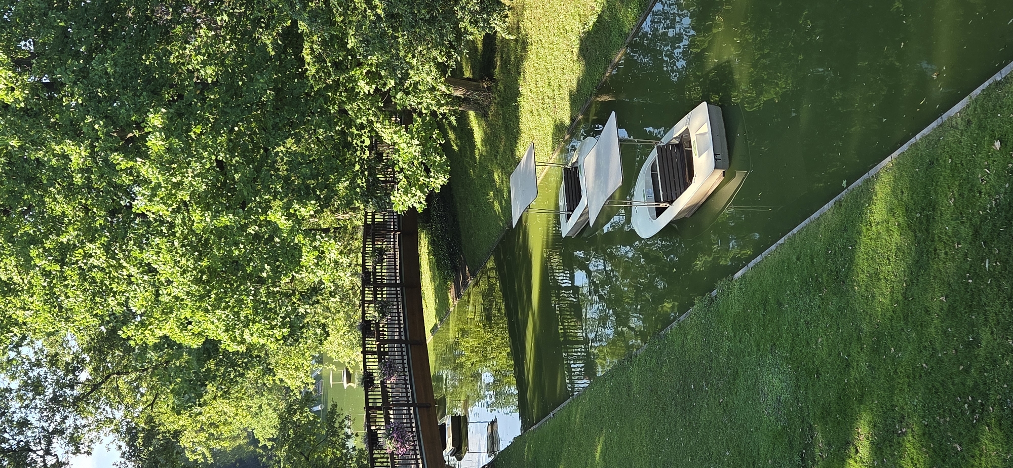 The image depicts a serene canal in the city of Karlsruhe, Germany, likely within the grounds of the Karlsruhe Zoo or its adjacent Hardtwald forest. The canal is bordered by lush green grass on both sides, with mature trees providing a verdant canopy overhead. A wooden bridge, adorned with flower planters, spans the canal in the middle ground, connecting the grassy banks.
In the canal, two small white pedal boats with beige canopies are visible. The boat closer to the foreground has the number "11" clearly marked on its stern. The water is a greenish hue, reflecting the surrounding foliage and the bridge. In the background, more of the canal and a distant body of water can be seen, also surrounded by trees. The lighting suggests a sunny day, with dappled sunlight falling on the grass and water. No people are present in the immediate scene.