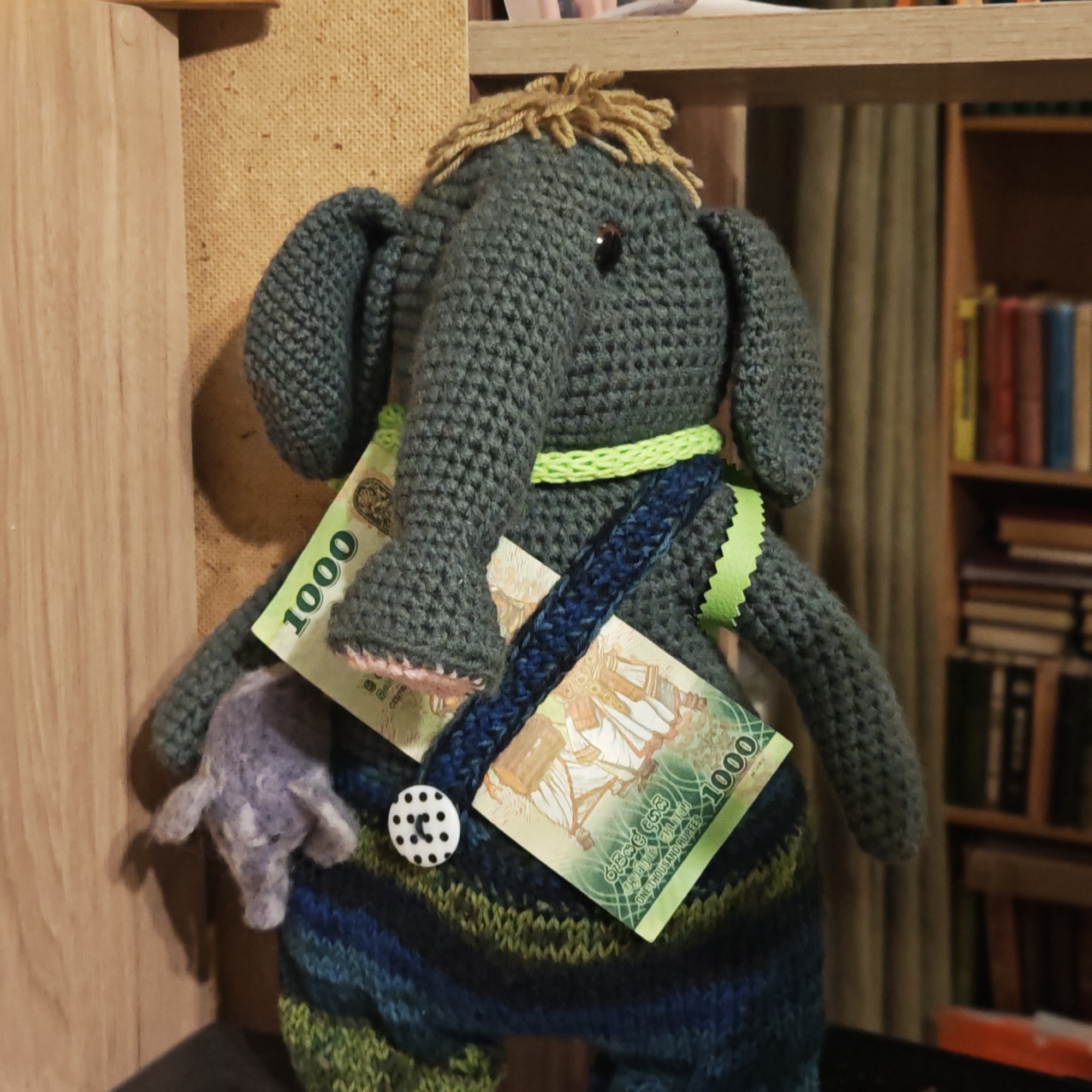 An eye-level shot captures a dark grey crocheted elephant plush toy, positioned in an indoor setting in Novo-Nikol'skoe, Russia. The elephant has light brown yarn "hair" on its head, small black bead eyes, and a hint of pink in its inner ears and snout. It wears a lime green crocheted band around its neck, a dark blue crocheted strap across its chest, and a ruffled lime green ribbon tied around its left side. Its lower body is crocheted in a striped pattern of blue, green, and black, adorned with a white polka-dotted button.
The plush toy's trunk holds a small, light purple, felted elephant figurine. Two Sri Lankan 1000 Rupee banknotes are tucked into its accessories. The text "1000", "ONE THOUSAND RUPEES", "CENTRAL BANK OF SRI LANKA", and "ශ්රී ලංකා මහ බැංකුව" (Central Bank of Sri Lanka in Sinhala) are clearly visible on the banknotes, along with intricate patterns and architectural illustrations.
The background consists of light wooden shelves or furniture on the left, and a beige, textured wall covering behind the plush. To the right, a wooden bookshelf filled with books of various colors and sizes is partially visible, suggesting a cozy, domestic environment. The lighting is soft, highlighting the textures of the crocheted toy and felted figurine.