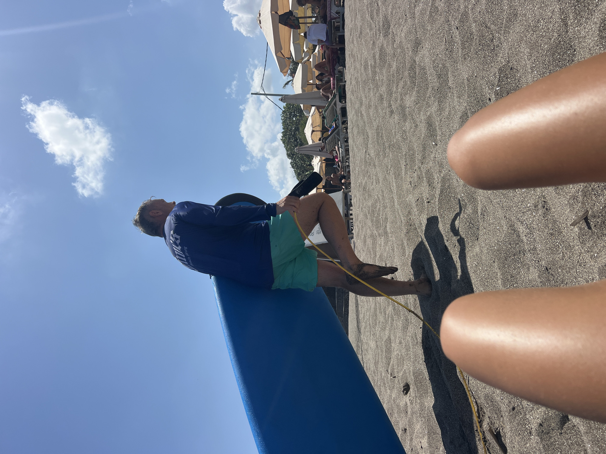 A man in a blue long-sleeved shirt and green shorts carries a large blue surfboard on a black sand beach in North Kuta, Indonesia. He is walking away from the camera, and his legs are covered in sand. In the background, beach umbrellas and lounge chairs are set up, with people relaxing. The sky is blue with a few white clouds. The perspective suggests the photo was taken from someone lying down on the sand, with their legs in the foreground.