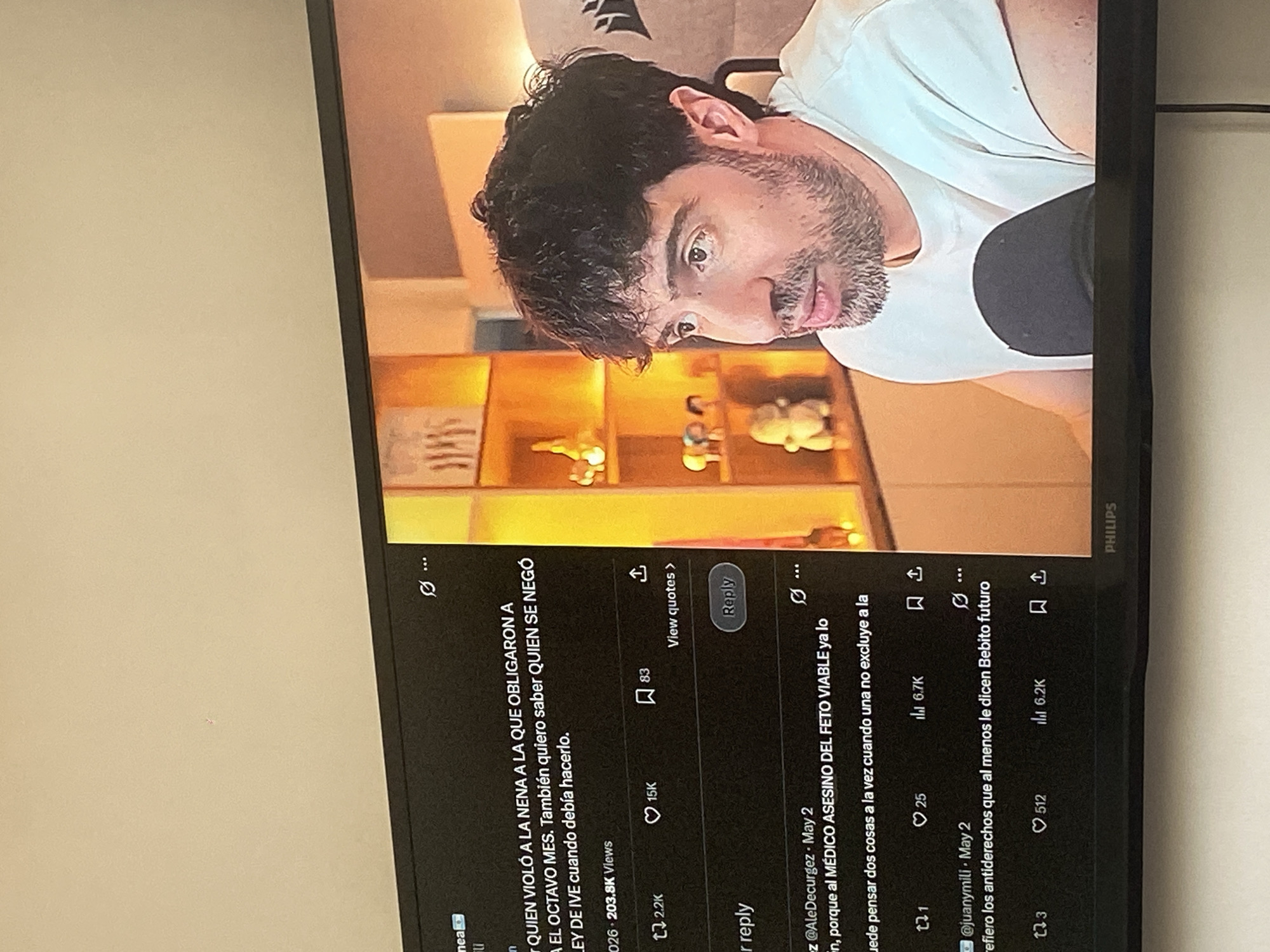 The image shows a close-up of a Philips television screen displaying a social media post, likely from Twitter, in Spanish. The post discusses a controversial issue involving violence against a minor and the denial of services related to the law of IVE (Interruption of Pregnancy). The screen also shows user comments discussing the matter, one of which mentions a "doctor assassin of the viable fetus."
In the upper part of the screen is a close-up of a man's face, likely the person whose social media content is being displayed. He has dark hair and a beard and is wearing a white shirt.
The location of the image capture is implied to be in Buenos Aires, Argentina, based on the language used in the social media posts and potential references to the "IVE" law, which is relevant in the Argentinian context. The setting appears to be indoors.
