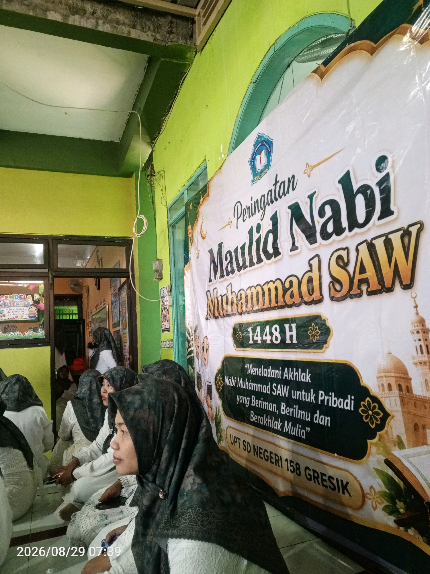 The image captures a group of women sitting on the floor, facing away from the camera and towards a banner. The banner, prominently displayed and partially obscuring some of the women, reads "Peringatan Maulid Nabi Muhammad SAW" and "1448 H". Below this, there is a quote in Indonesian that translates to "Emulating the Morals of Prophet Muhammad SAW for a Pious, Knowledgeable, and Virtuous Self." The banner also indicates "UPT SD NEGERI 158 GRESIK," suggesting the event is hosted by a state elementary school in Gresik. The women are dressed in modest attire, with most wearing hijabs in dark colors and white clothing. The environment appears to be an indoor space with bright green walls. A window is visible to the left of the banner, showing a glimpse of another room. The timestamp "2026/08/29 07:39" is overlaid on the bottom left corner of the image, which is likely a date and time stamp from the camera. The overall scene depicts a religious gathering or commemoration event.