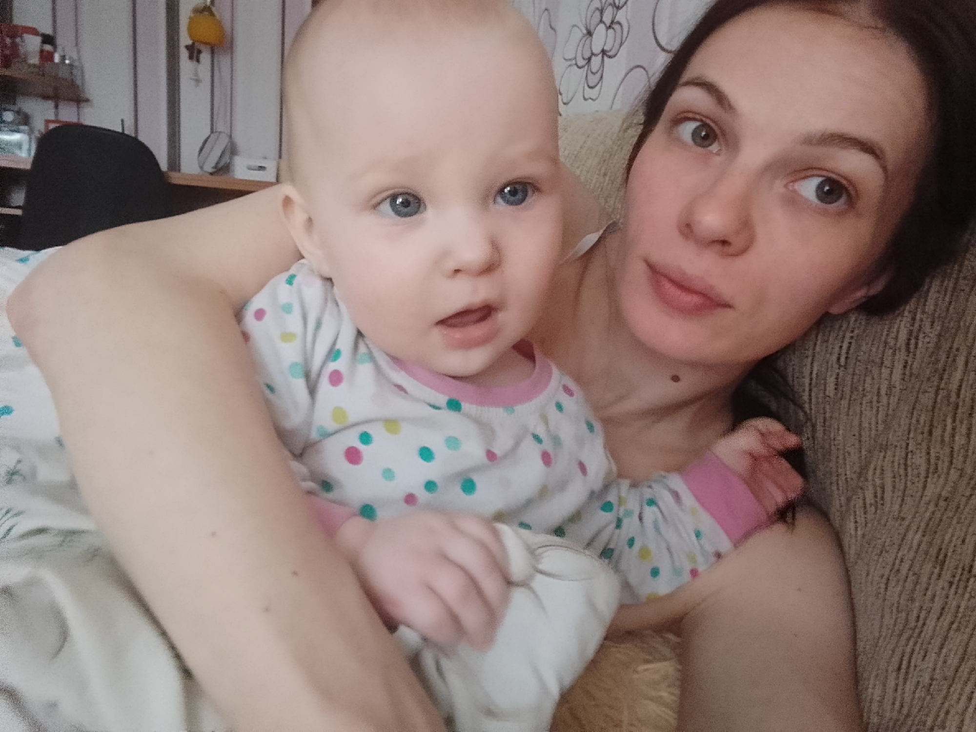 The image is a selfie taken indoors. In the foreground, a woman is holding a baby. The woman is on the right side of the frame, and the baby is on the left. The baby is facing the camera with wide blue eyes and an open mouth. The woman is also looking at the camera. Both are wearing casual clothing. The background is blurred, but it appears to be a room with patterned wallpaper and furniture. The scene suggests a domestic setting, likely a home in Minsk, Belarus.