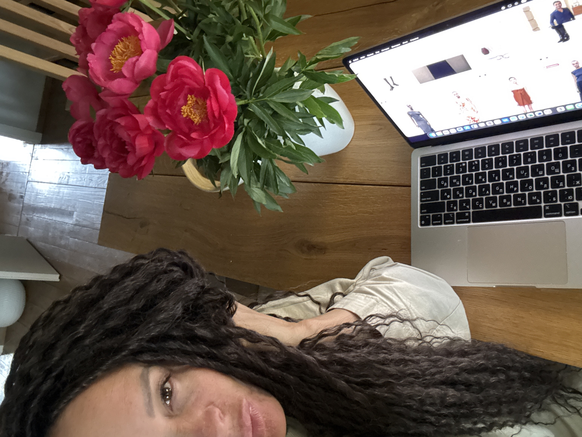 The image presents a top-down view of a person lying on a wooden surface, engaged with a laptop, alongside a vibrant floral arrangement. The main subject is a woman with long, dark, curly hair, whose face – including her left eye, nose, and lips – is partially visible as she looks towards the camera. She is wearing a light-colored garment, possibly a beige or off-white shirt.
Adjacent to her, a silver laptop (resembling a MacBook) is open, its screen displaying what appears to be an e-commerce website or a digital mood board. The screen shows various images of models presenting different clothing items and accessories. A notable detail is the laptop's keyboard, which clearly features Cyrillic characters alongside Latin ones, indicating a Russian layout.
Between the person and the laptop lies a beautiful bouquet of pink peonies with striking yellow centers, surrounded by rich green leaves, held in a simple vase. The surface beneath them is a warm-toned wooden table. In the blurred background, hints of other indoor elements like wooden slats and a white, rounded object can be seen.
The scene suggests an indoor setting, likely a home, where the individual is either working or relaxing with the laptop. The angle implies a selfie or an overhead shot. The Cyrillic keyboard characters are a strong indicator of the geographical context, consistent with the provided location of Chaikovsky, Russia. The person's expression is neutral, adding to the relaxed atmosphere.