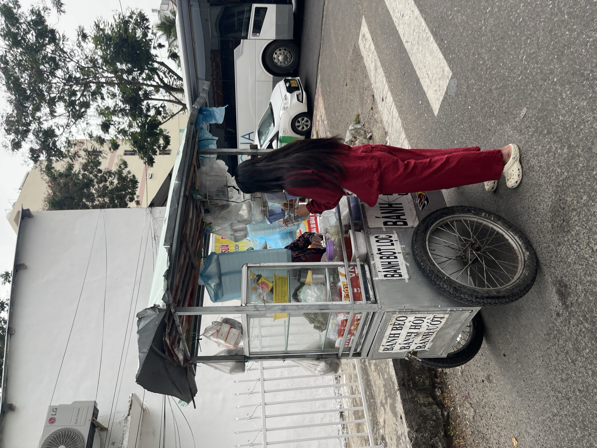 A street scene in Bac Nha Trang Ward, Vietnam shows a food vendor, a woman with long black hair dressed in a red outfit and wearing white shoes, interacting with a food cart. The cart has a variety of food items displayed behind clear panels and the Vietnamese words "Bánh Bèo, Bánh Hỏi, Bánh Ướt, Bánh Bột Lọc" are printed on its side. The cart is situated near a crosswalk, with a white bus and a car parked nearby. An LG air conditioning unit is visible on a building to the left of the cart. A tree and a building are in the background. The day is overcast.