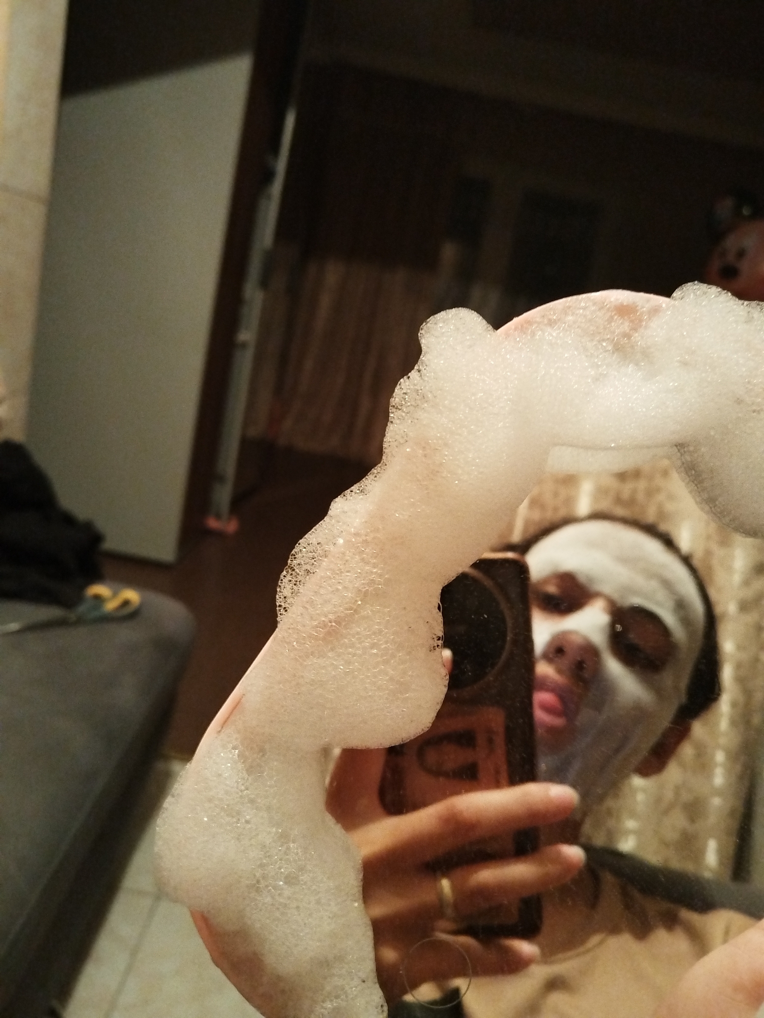 A person is taking a selfie in a bathroom mirror. The person is holding a cell phone in their left hand. They are wearing a white facial mask. The person is holding a handful of white foam. The background of the image shows part of a gray sofa and a white door.