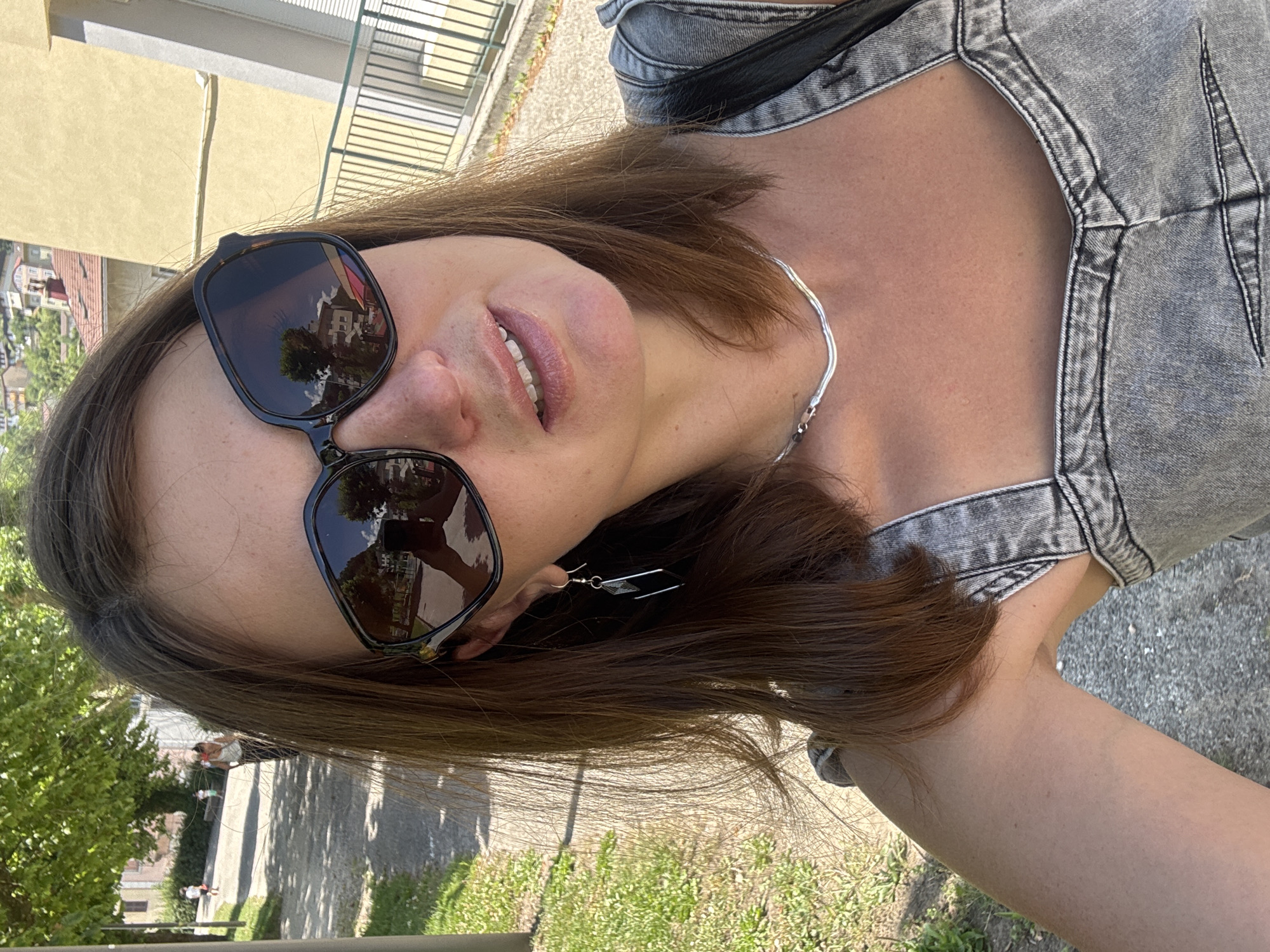 A woman is taking a selfie outdoors on a sunny day. She has brown hair and is wearing sunglasses, a silver necklace, and a denim top. In the background, there is a tree-lined path with people walking, and a building with yellow walls. The location is Saint-Claude, France.