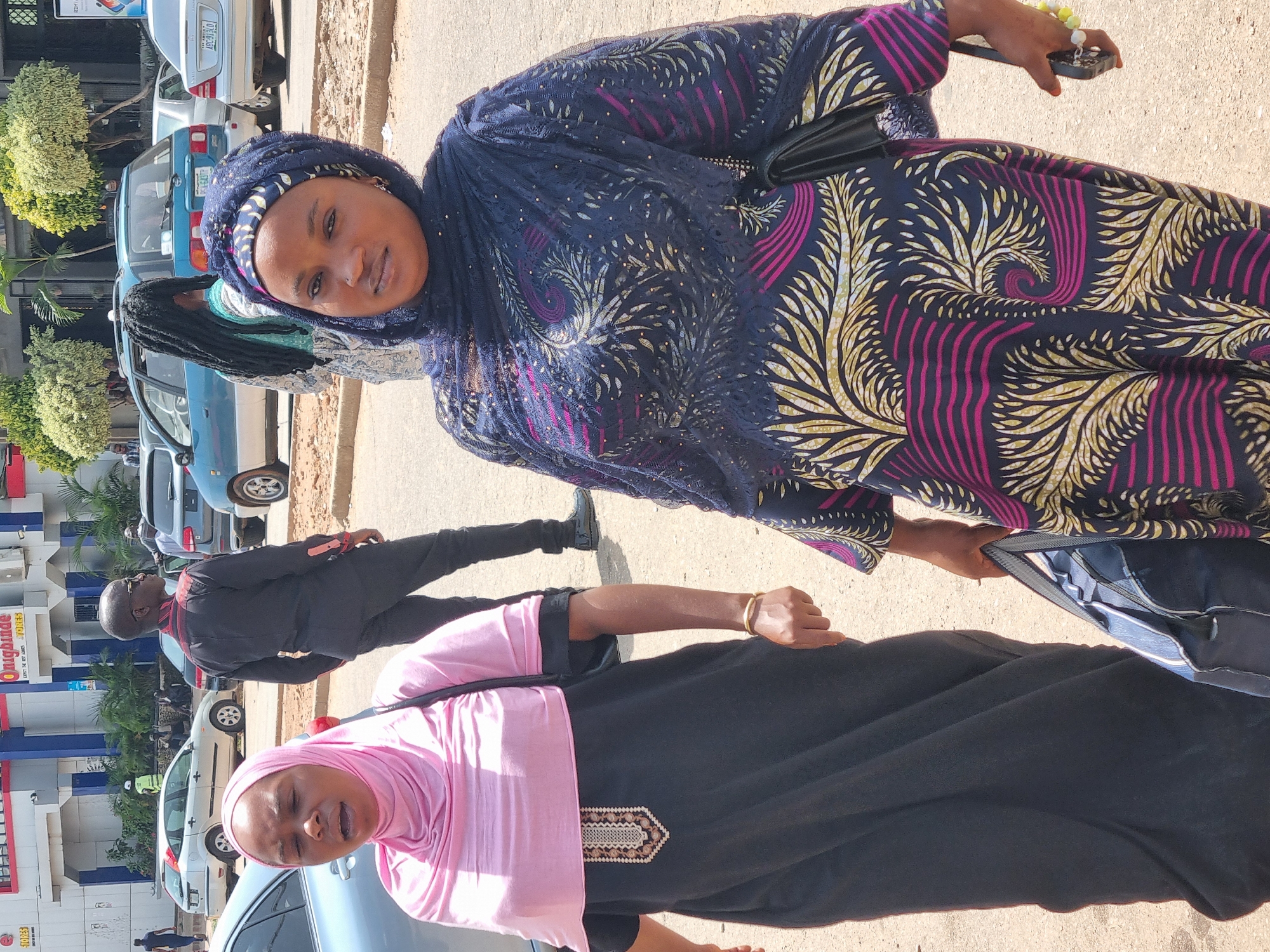 In Jos, Nigeria, two women are seen walking on a paved road near a building with red and white signage. The woman on the left wears a pink hijab and a black dress with an embroidered chest, carrying a black bag over her shoulder. She is mid-conversation, with her mouth open. The woman on the right wears a dark blue hijab and a colorful dress with a swirling pattern of dark blue, pink, and yellow. She holds a phone in her right hand and has a black bag slung over her shoulder. In the background, several cars are parked or moving, including a silver sedan, a blue hatchback, and other vehicles. A man in a dark suit with sunglasses stands to the left of the women, facing away from the camera. The environment appears to be an urban setting with trees and buildings visible.