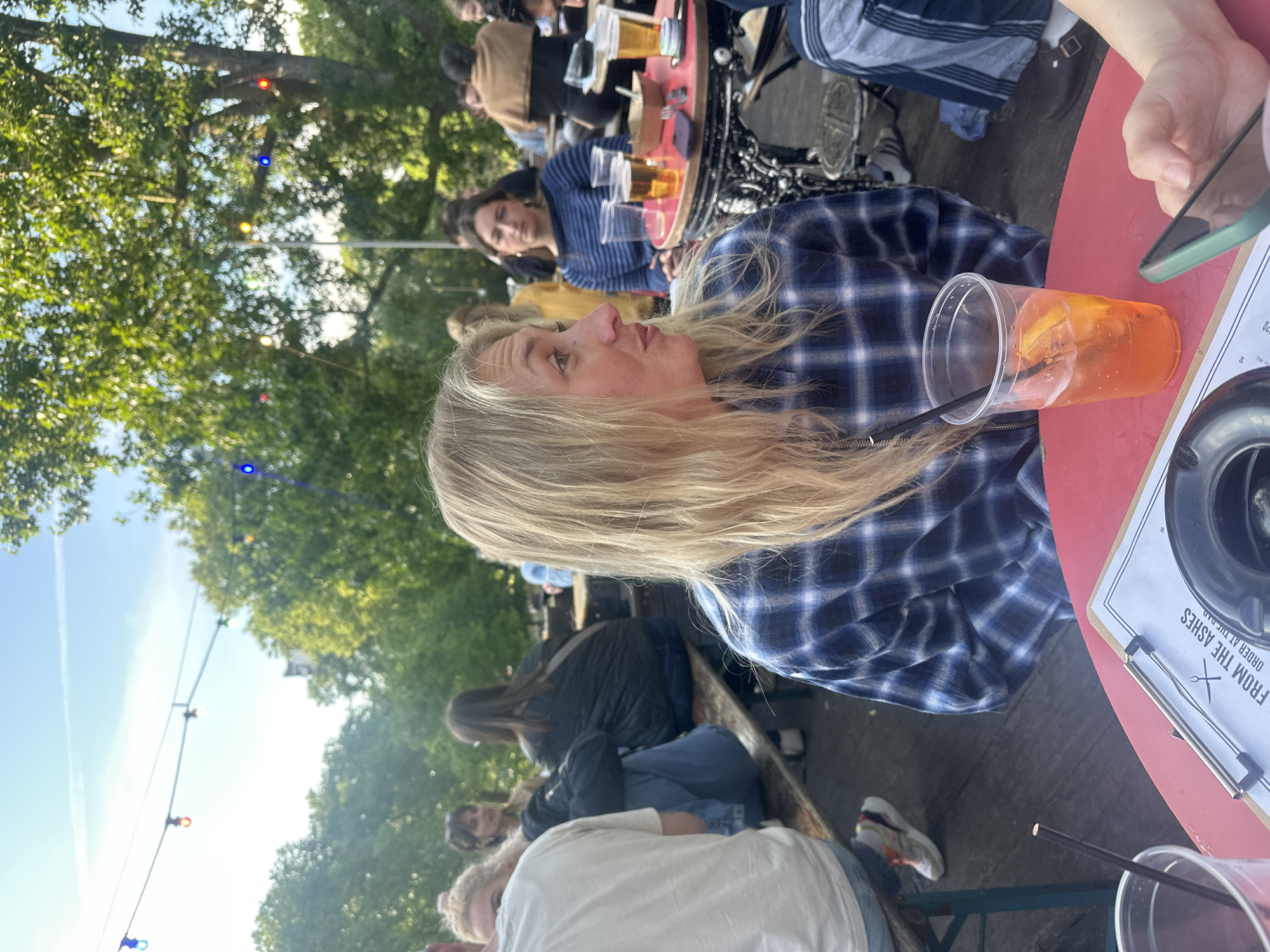 The image shows a group of people outdoors, likely at a bar or restaurant. A woman with blonde hair wearing a blue and white plaid shirt is seated at a red table with a plastic cup filled with an orange beverage. A menu board is visible on the table. Other people are seated at tables in the background, some with drinks. The scene appears to be in an urban outdoor setting, with trees and string lights visible overhead. It is likely taken in London, United Kingdom, based on the bar’s name on the menu board.