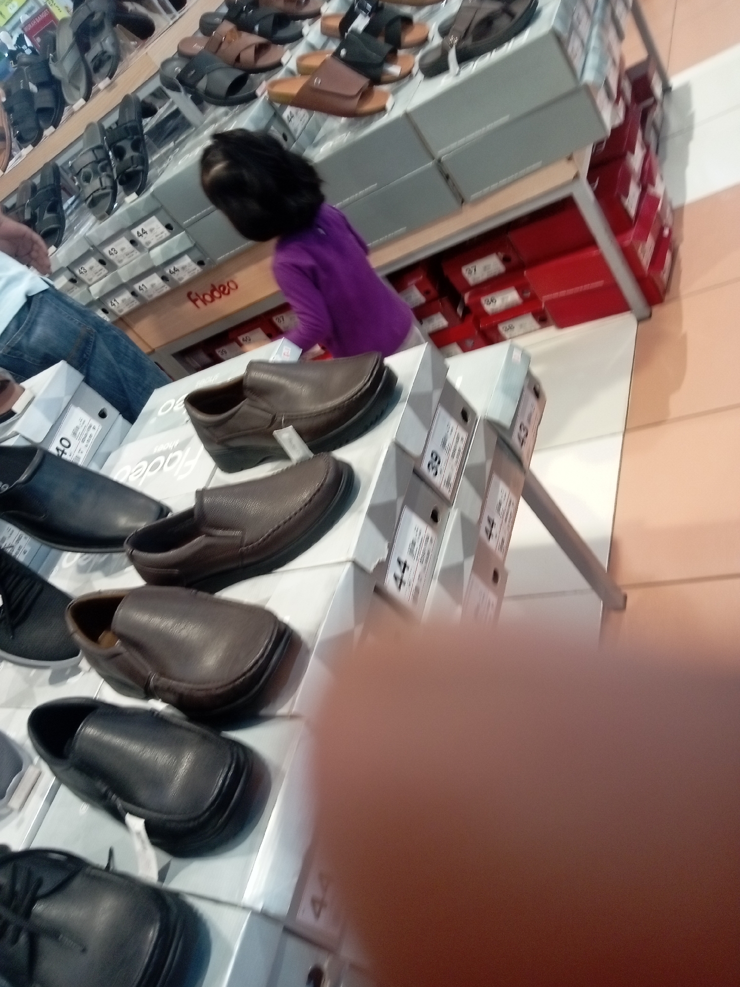 The image depicts a retail environment, likely a shoe store, with several racks of shoes and shoe boxes. In the foreground, a child with dark hair is wearing a purple long-sleeved shirt and is positioned to the left of center, appearing to be looking at the shoes. Several pairs of men's dress shoes and loafers are visible on display, primarily in brown and black leather. Some shoes are placed on top of boxes, while others are inside or on shelves. The background shows rows of shoe boxes, some red and some gray, with visible labels and size markings. The overall impression is a well-stocked shoe retail section within a store. Based on common retail layouts and the style of merchandise, this scene is consistent with locations found in shopping malls in Batam, Indonesia, such as Mega Mall or BCS Mall.