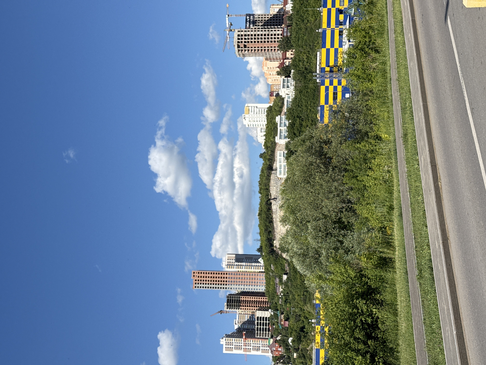 The image shows an urban landscape with several tall buildings under construction, set against a bright blue sky with scattered white clouds. The buildings are a mix of modern apartment blocks and office structures, some of which are in various stages of completion, with visible scaffolding and cranes. The scene appears to be in a developing area, as indicated by the construction activity.
To the right of the buildings, there is a lush green slope covered in trees and bushes. Beyond this green area, a paved road runs parallel, with concrete barriers and grassy verges separating it from the slope. On the far right of the image, the road surface and markings are visible, suggesting it is a modern, well-maintained route.
There are prominent blue and yellow striped structures along the edge of the green slope, which might be temporary fencing or signage related to the construction or a nearby event. One of the construction cranes has a sign attached that reads "HART 85-77477".
The lighting suggests it is daytime, likely midday or early afternoon, given the bright sunlight and clear sky. The weather appears to be fair and sunny, contributing to the overall cheerful and optimistic atmosphere of urban development and growth. The orientation of the image is vertical, meaning the scene is likely captured with the camera held upright.