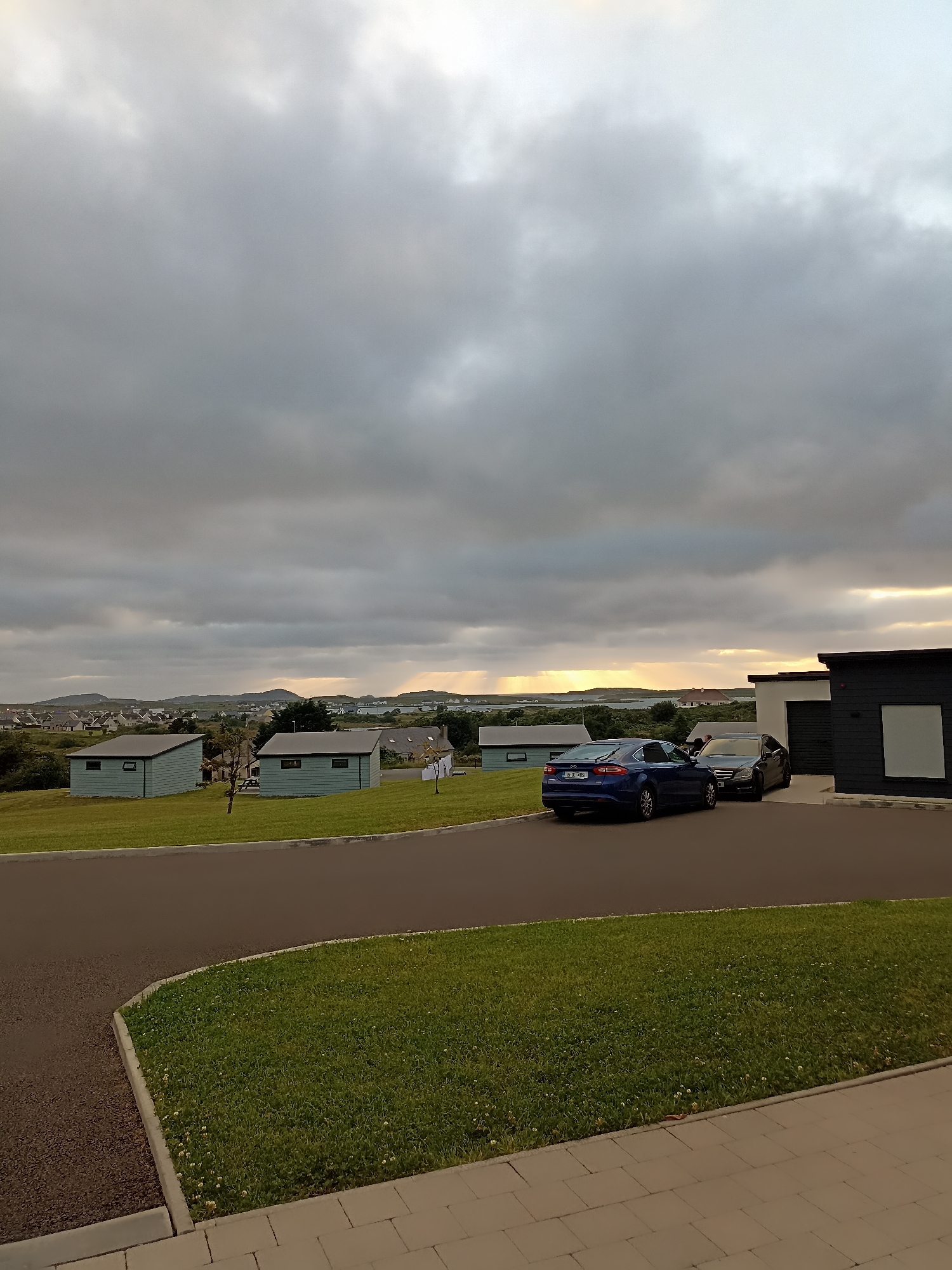 This image depicts a scene in Doirí Beaga, Ireland, with a focus on a residential area during what appears to be late afternoon or early evening. The sky is heavily overcast with dramatic, grey clouds, but a sliver of sunlight is breaking through near the horizon, casting a warm glow on distant hills and water.
In the foreground, a paved driveway curves towards the viewer, bordered by a well-maintained lawn. To the right, a modern, dark-colored building with a light-colored section and what appears to be a garage door is visible. Two cars are parked on the driveway: a dark-colored sedan and a blue sedan in front of it.
Beyond the immediate foreground, several light-blue, rectangular structures with dark roofs are arranged on a grassy slope. These structures resemble small houses or cabins. In the background, the landscape reveals a dispersed collection of houses with white and light-colored roofs, typical of a rural Irish setting. Rolling hills and bodies of water are visible in the far distance, bathed in the soft light from the setting sun. The overall impression is one of a serene, albeit cloudy, evening in a picturesque Irish locality.