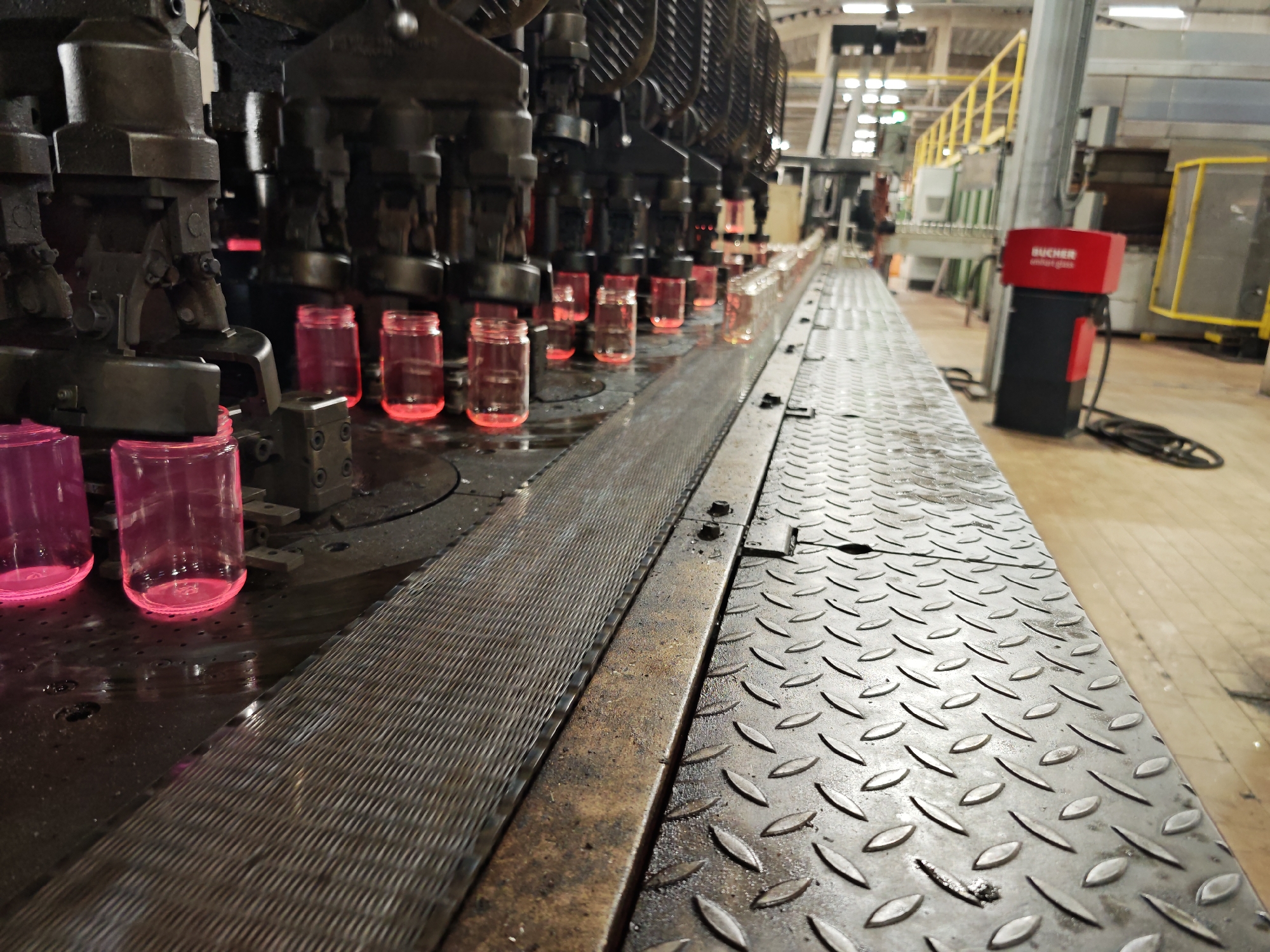 The image depicts an industrial setting, possibly a glass manufacturing facility. Rows of glass jars, with a reddish glow suggesting they are still hot, are being processed on a conveyor belt system. Automated machinery, consisting of dark, heavy mechanical units, is positioned above the jars, presumably part of the production or cooling process. The conveyor belt is made of a metal surface. In the background, there are other machines and industrial equipment, and a red machine labeled "BUCHER" is visible. The floor is light-colored and shows signs of industrial use.
There is no verifiable information to suggest the location in the city of Pleven, Bulgaria.