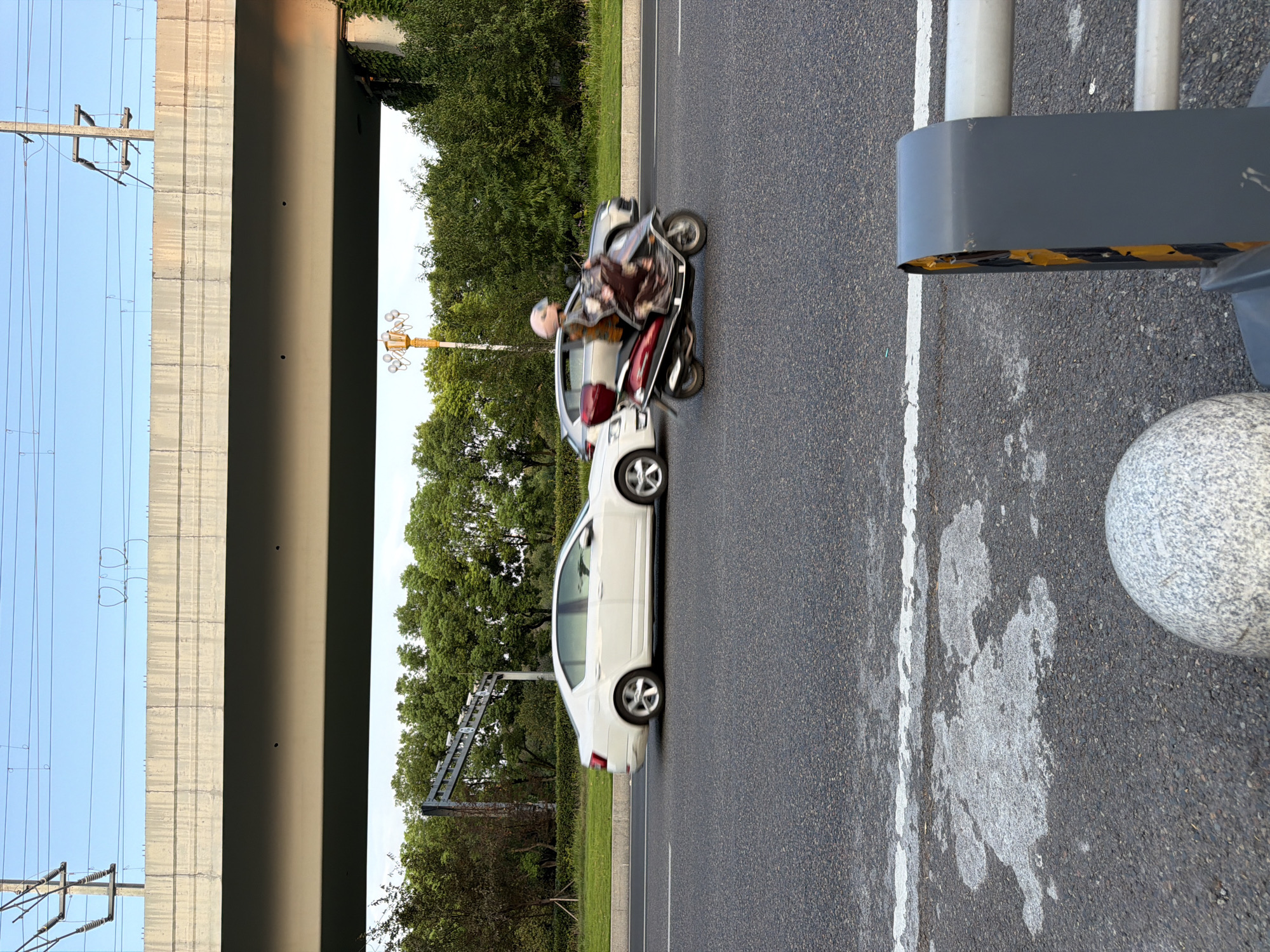 In Jiangjin, China, a red and silver scooter carrying a person wearing a pink helmet and a patterned outfit is moving on a road under an elevated highway. The scooter is to the right of a white sedan. The road is asphalt, and there are white lane markings. Trees and vegetation are visible in the background, and overhead, power lines and the concrete structure of the highway are present. In the foreground, a section of a gray and yellow traffic barrier and a spherical gray object are visible. The lighting suggests it is either early morning or late afternoon.