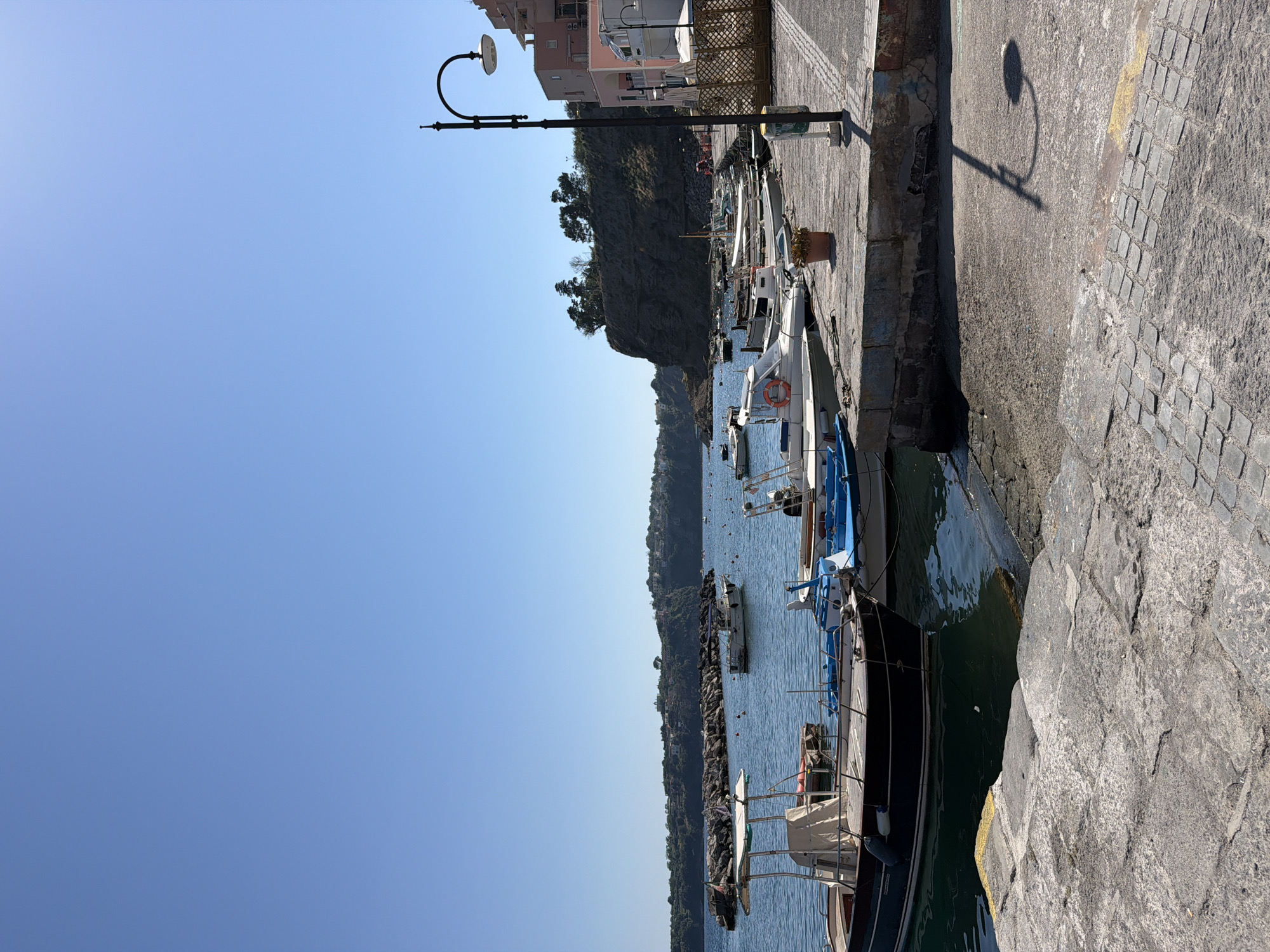 The image shows a harbor scene in Procida, Italy. The view is of a row of fishing boats moored along a concrete quay. A street lamp is visible along the quay, casting a shadow onto the pavement. Buildings are visible behind the quay with a cliff and trees. The sea is calm and blue.