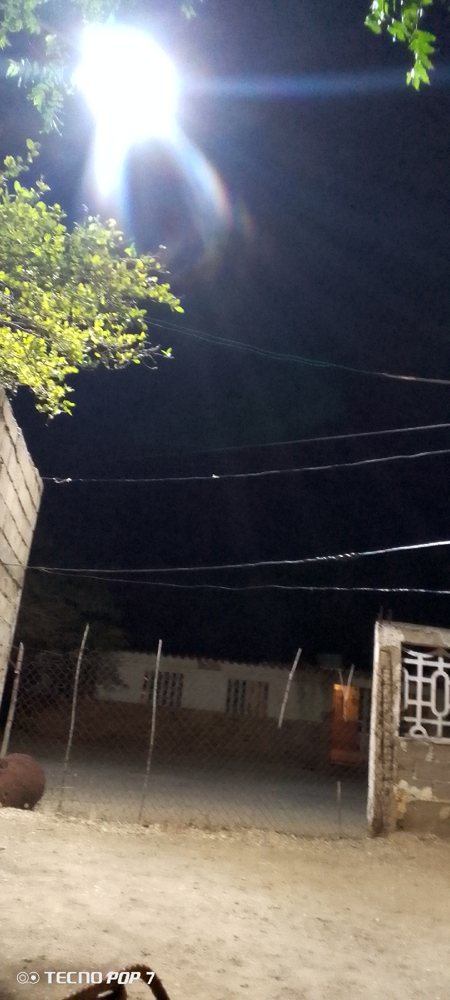 The image shows a nighttime view in what appears to be a residential area. Bright light obscures the upper left of the frame, possibly a streetlight or the moon. Overhead are power lines spanning the scene. A chain-link fence runs across the midground, behind which is a low building with windows. An object resembling a light fixture is visible near the right edge of the fence. The ground is light-colored, potentially dirt or sand. A concrete structure is present on either side of the frame on the lower left and right corners. The location is Altagracia, Venezuela. No people are visible in the scene.