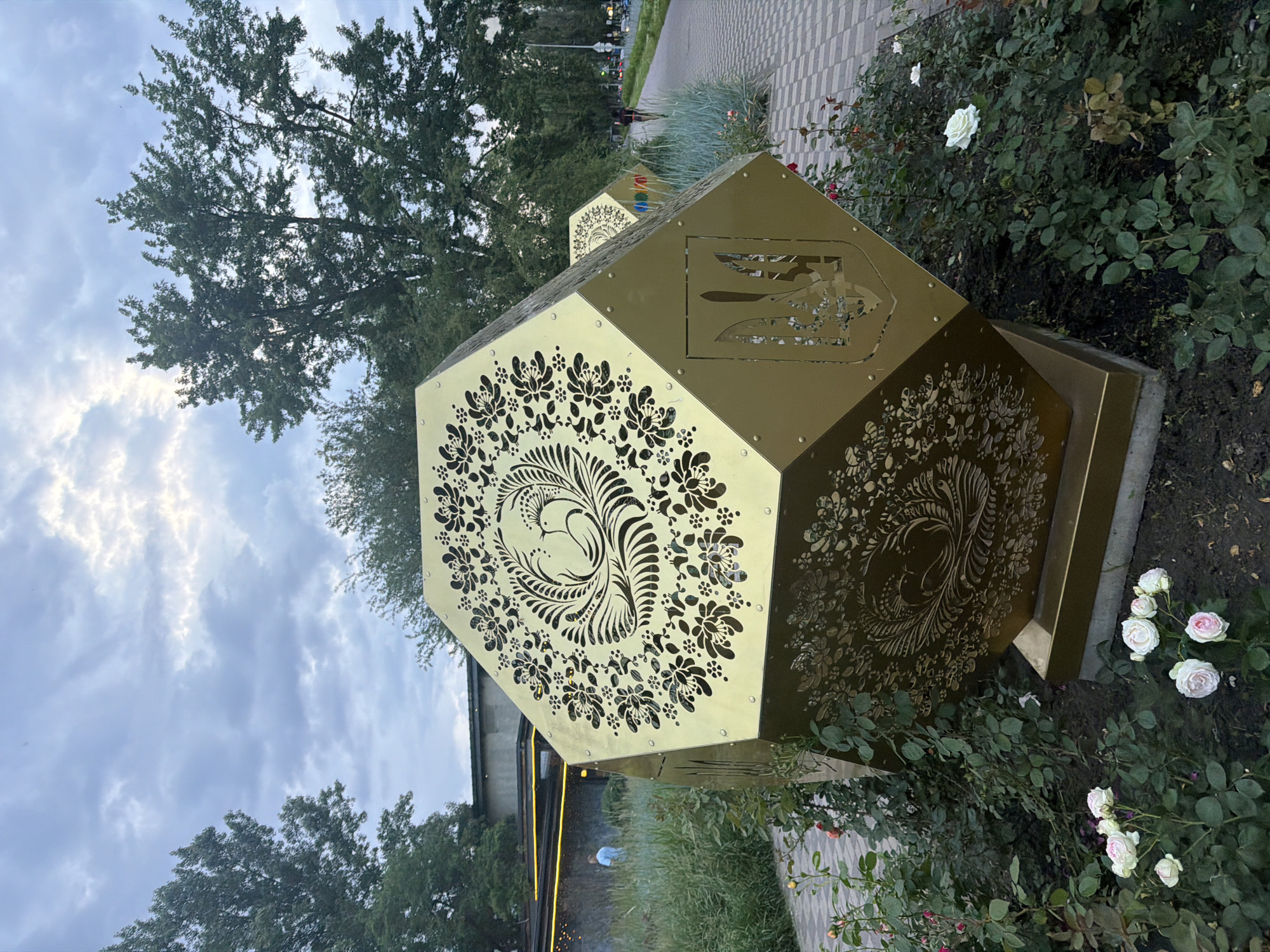 The image depicts a large, faceted, golden sculpture adorned with intricate floral patterns and national symbols, likely the Ukrainian Tryzub (trident). The sculpture is situated outdoors, surrounded by a flower bed of roses, with a paved walkway and greenery in the background. In the distance, trees line the walkway, and a building with yellow lighting is visible. The sky is overcast with clouds. The sculpture is located in the city of Kyiv, Ukraine. The scene is a public space, possibly a park or a street, and appears to be during daylight hours, although the lighting suggests it might be late afternoon or early evening. There are no discernible actions or interactions occurring.