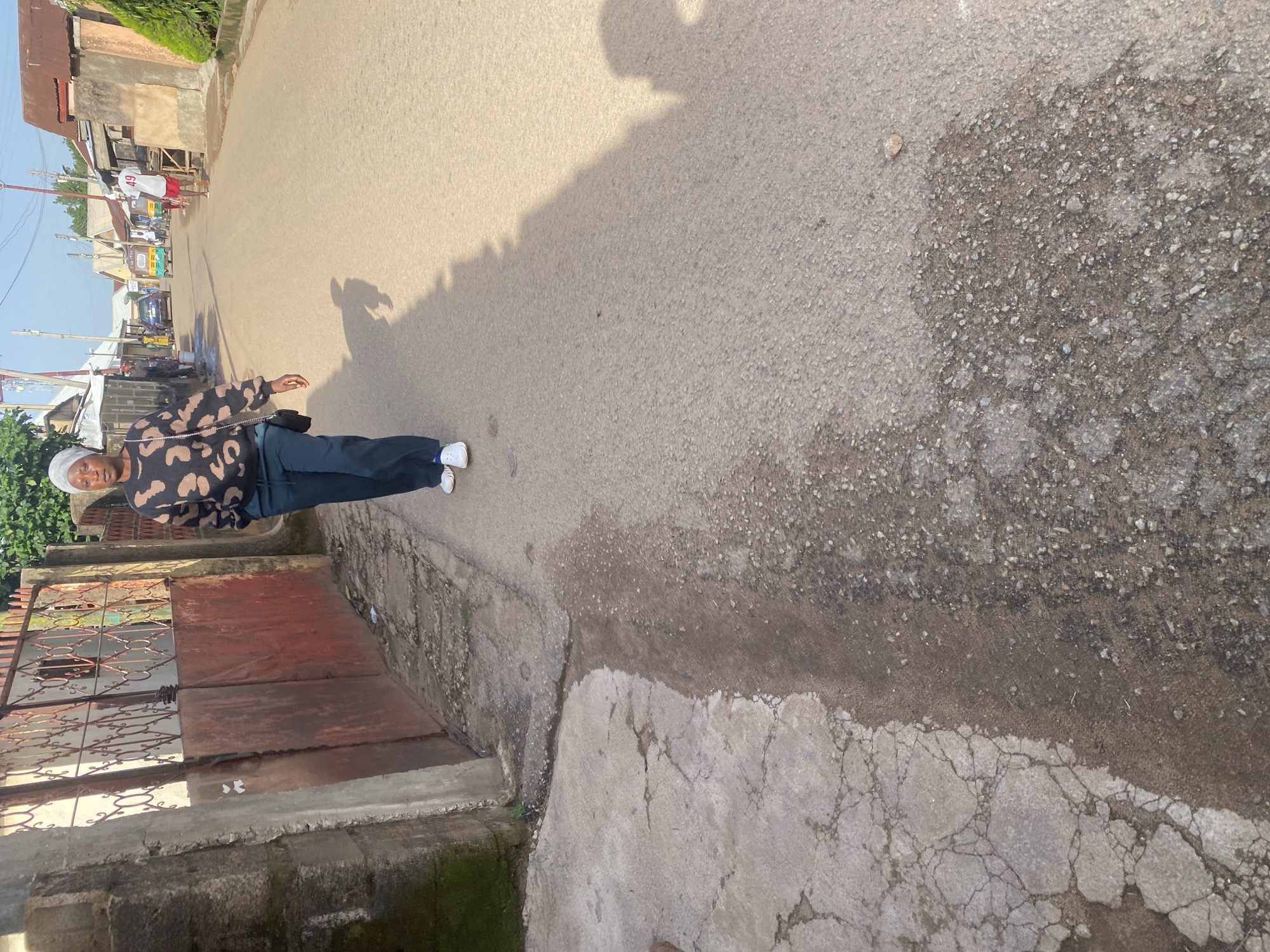 A person wearing a white headscarf, a dark sweater with a light-colored abstract pattern, and dark pants stands on a cracked asphalt road. White sneakers are visible on their feet. The person is partially silhouetted against a bright, hazy sky. Behind them, a street scene unfolds with buildings, some with corrugated metal roofs, and several vehicles, including a tuk-tuk. The sunlight casts a long shadow of the person onto the road. The atmosphere suggests it is daytime, possibly late afternoon, given the long shadow. The environment appears to be an urban or semi-urban area, with elements of everyday life, such as traffic and structures, visible. The overall impression is of a candid street photograph capturing a moment in time within a Nigerian setting.