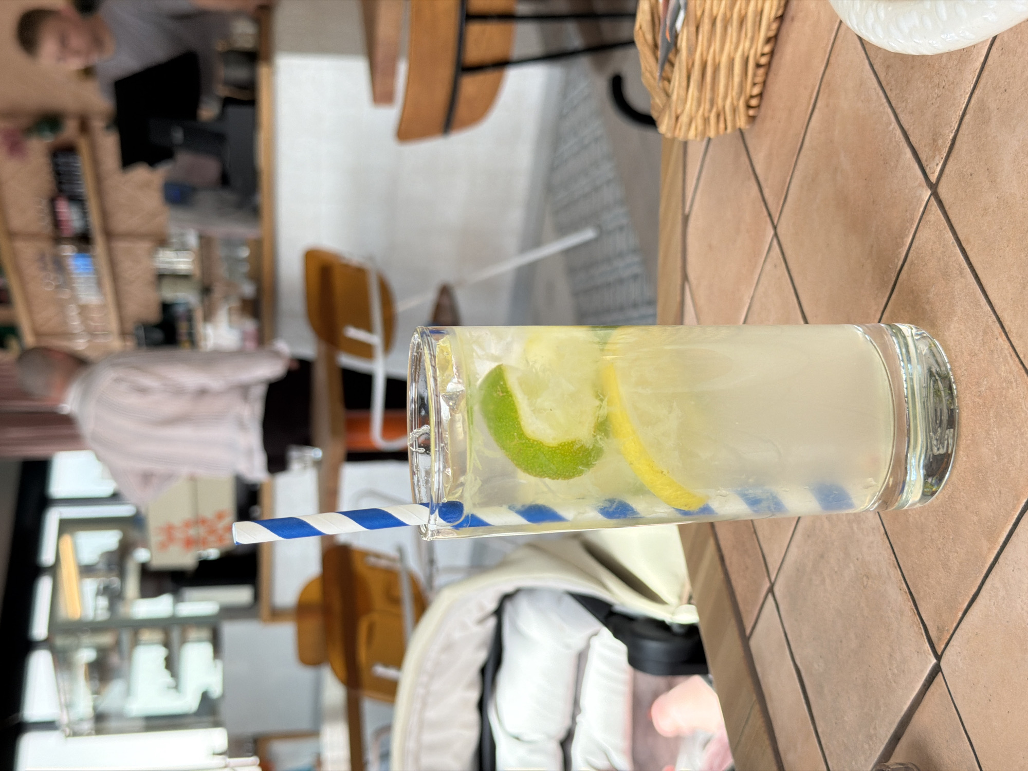 The image shows a glass of lemonade with a blue and white striped straw on a brown tile table. The lemonade contains slices of lime and lemon. In the background, there are glimpses of a restaurant interior with blurred figures, chairs, and a bar area. The image does not contain sufficient information to identify the specific location in Moscow, Russia, or provide any additional details about the city.