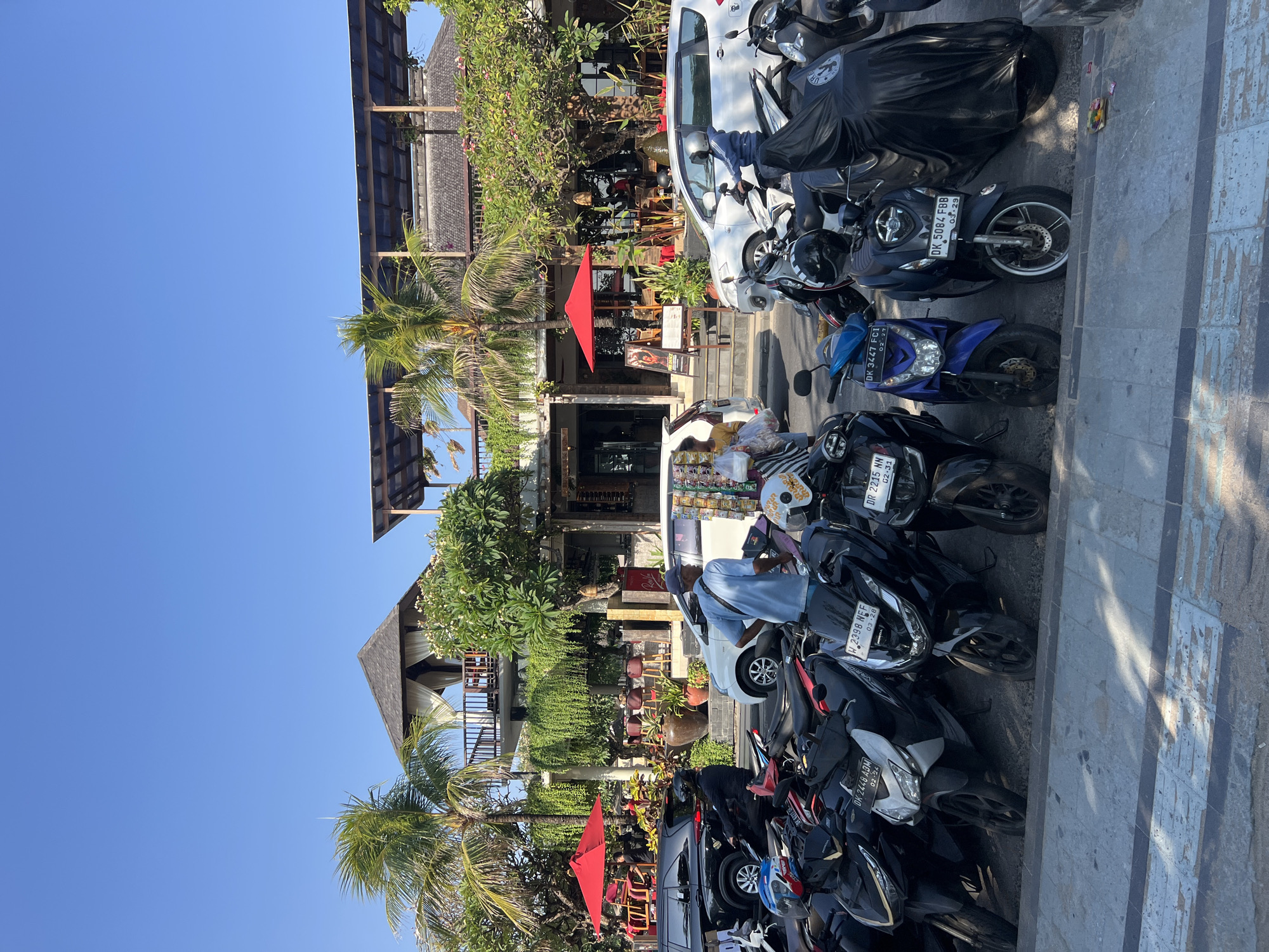 This is a vertical, eye-level, medium shot photograph taken in Kuta, Indonesia, on a clear, sunny day. The scene depicts a street with a building on the right and a row of parked motorcycles in the foreground.
The building is a multi-story structure with a Balinese architectural style, featuring a thatched roof and balconies adorned with greenery. A prominent thatched-roof pavilion sits on the upper level, with white curtains visible. Several palm trees surround the building. Red umbrellas are set up outside the establishment.
In front of the building, a variety of motorcycles are parked in a row. Some are covered with black tarps, while others are exposed. A few people are visible, including a man in a blue shirt standing by a motorcycle, and a woman in a yellow shirt walking near a white car. There are also two cars parked on the street, one black and one white.
The foreground shows a sidewalk and the street where the motorcycles are parked. The overall impression is of a busy commercial area in a tropical location.