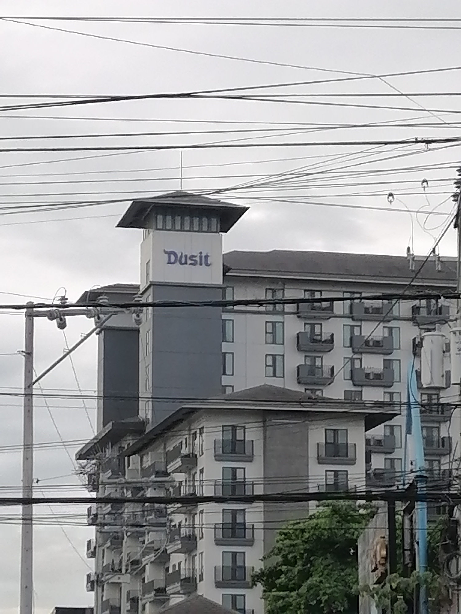 The image shows a tall building with the "Dusit" logo visible on its upper section. The building appears to be a hotel or condominium complex, characterized by its multi-story structure with numerous balconies and windows. The surrounding environment is urban, with visible utility poles and numerous overhead wires crisscrossing the sky, obscuring parts of the building and the sky. The lighting suggests an overcast day. The presence of the "Dusit" branding indicates this is likely a Dusit branded establishment, and given the context of Davao City, Philippines, this could be Dusit Thani Residence Davao or a similar property of the Dusit International hotel group in the area.