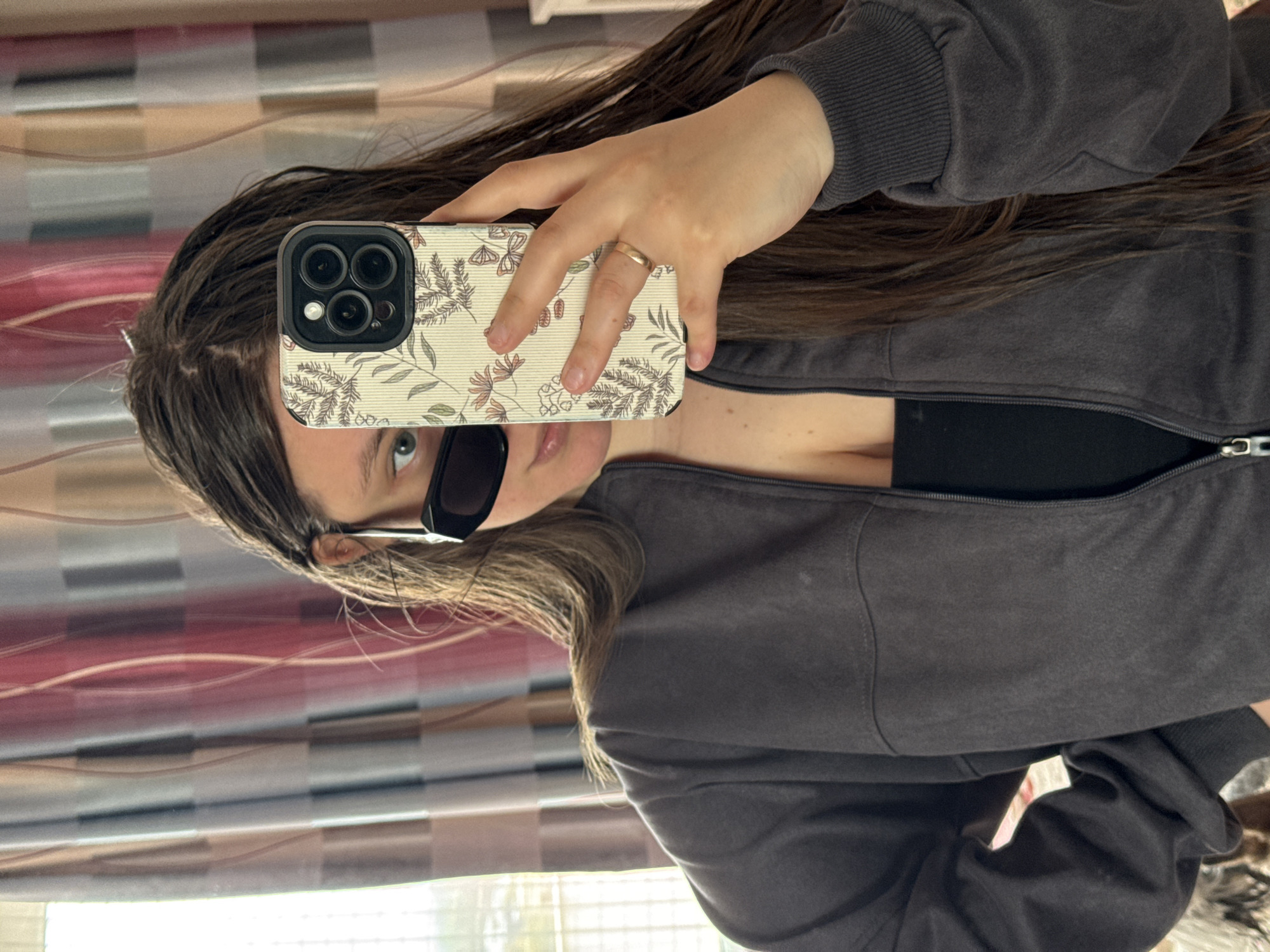 A woman takes a selfie with a smartphone in front of a vertically-oriented window. She holds the phone with her right hand, her hand is displaying a wedding band. She wears a gray zip-up jacket over a black top and sunglasses. Her hair is brown. Behind her is a window with horizontally oriented blinds. There is no evidence of the location being in Krasnodar, Russia.