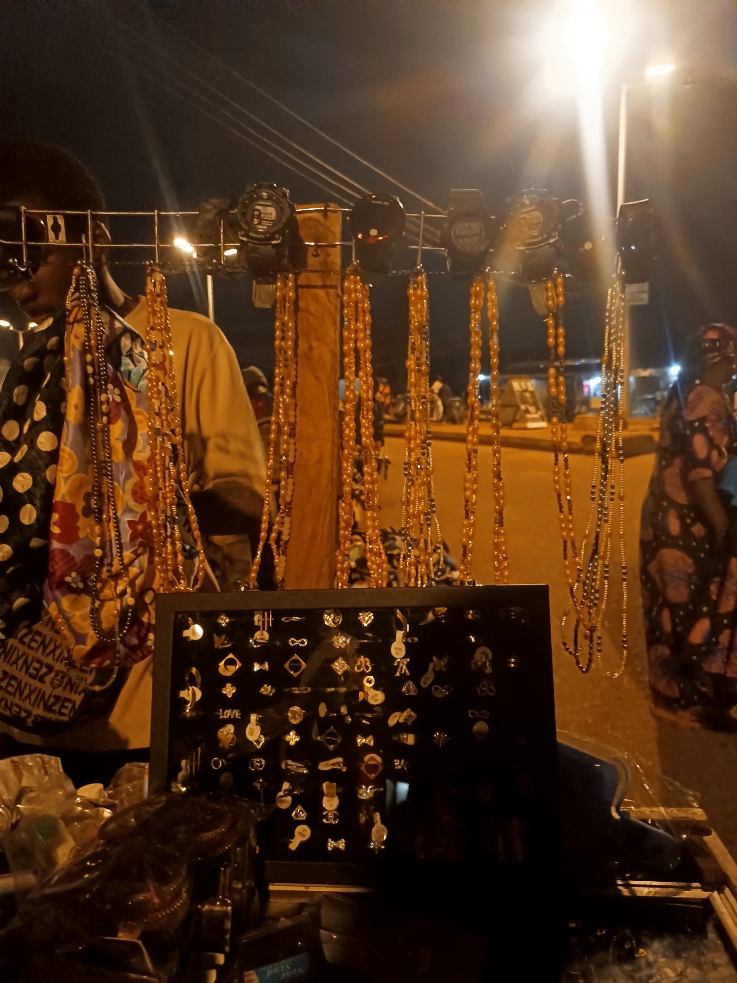 A street vendor displays merchandise at night in Bukuru, Nigeria. The goods include watches hanging from a metal grid, strands of amber and gold-toned beaded necklaces, and a display case filled with earrings. A man stands behind the merchandise display, wearing a yellow shirt and a patterned scarf. A woman in a patterned dress walks past in the background. A street light illuminates the scene and the surrounding road. Other people and vehicles are visible in the background.