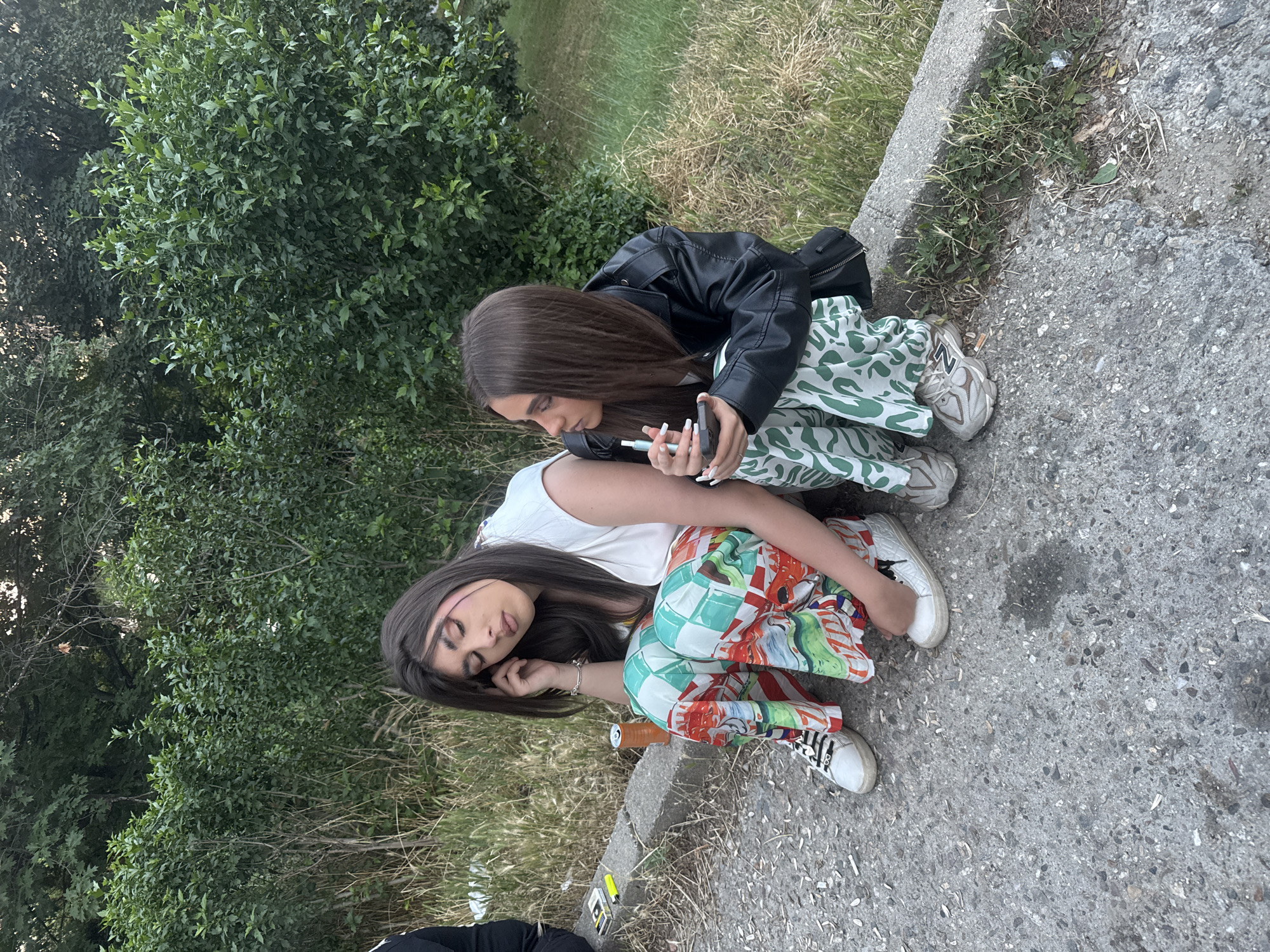 Two young women are sitting on a concrete curb next to a road, with a dense green hedge and trees in the background. The woman on the left has her eyes closed and is resting her head on her hand. She is wearing a white sleeveless top and brightly patterned wide-leg pants, with white sneakers. An orange can and a pack of cigarettes are on the ground near her. The woman on the right is looking towards the left and holding a vape pen and a smartphone. She is wearing a black leather jacket over a white top and patterned pants, with beige sneakers. The scene appears to be in an outdoor urban setting, possibly in Blagoevgrad, Bulgaria, given the context.