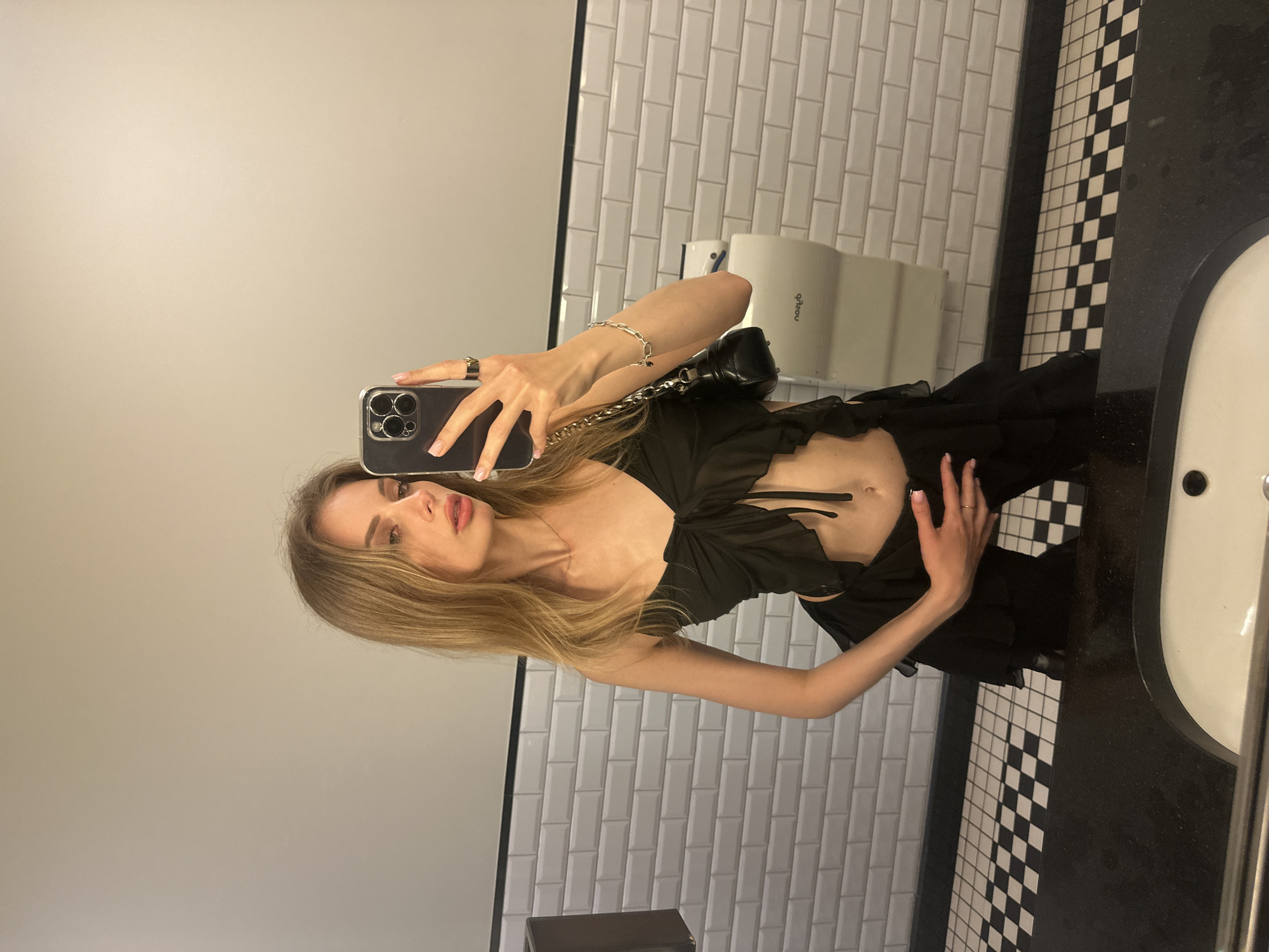 A young woman is captured in a mirror selfie within a public restroom, stated to be in Warsaw, Poland. She stands in a three-quarter pose, looking towards her smartphone held in her right hand, which captures her reflection. Her left hand rests on her lower abdomen. She is a slender individual with long, straight light brown hair and light makeup. She wears a black, sheer, ruffled crop top tied in the front, exposing her midriff and navel. Her lower garment consists of matching black, ruffled fabric. Accessories include a dark-colored smartphone, a dark ring on her right index finger, a thin gold-colored ring on her left ring finger, a silver chain-link bracelet on her left wrist, and a small black shoulder bag with a silver chain strap hanging over her left arm. Her fingernails are manicured with a light base and white tips.
The restroom features a lower wall section clad in white subway tiles with dark grout, topped by a light grey plain wall and a thin black horizontal border line. The floor is covered with black and white checkered tiles laid diagonally. A black, reflective countertop is prominent in the foreground, with a white oval sink and a black drain partially visible. A white hand dryer, with "Dyson" reflected as "nozyD", is mounted on the tiled wall to the right of the woman. A dark-colored dispenser is partially visible on the left side of the frame.