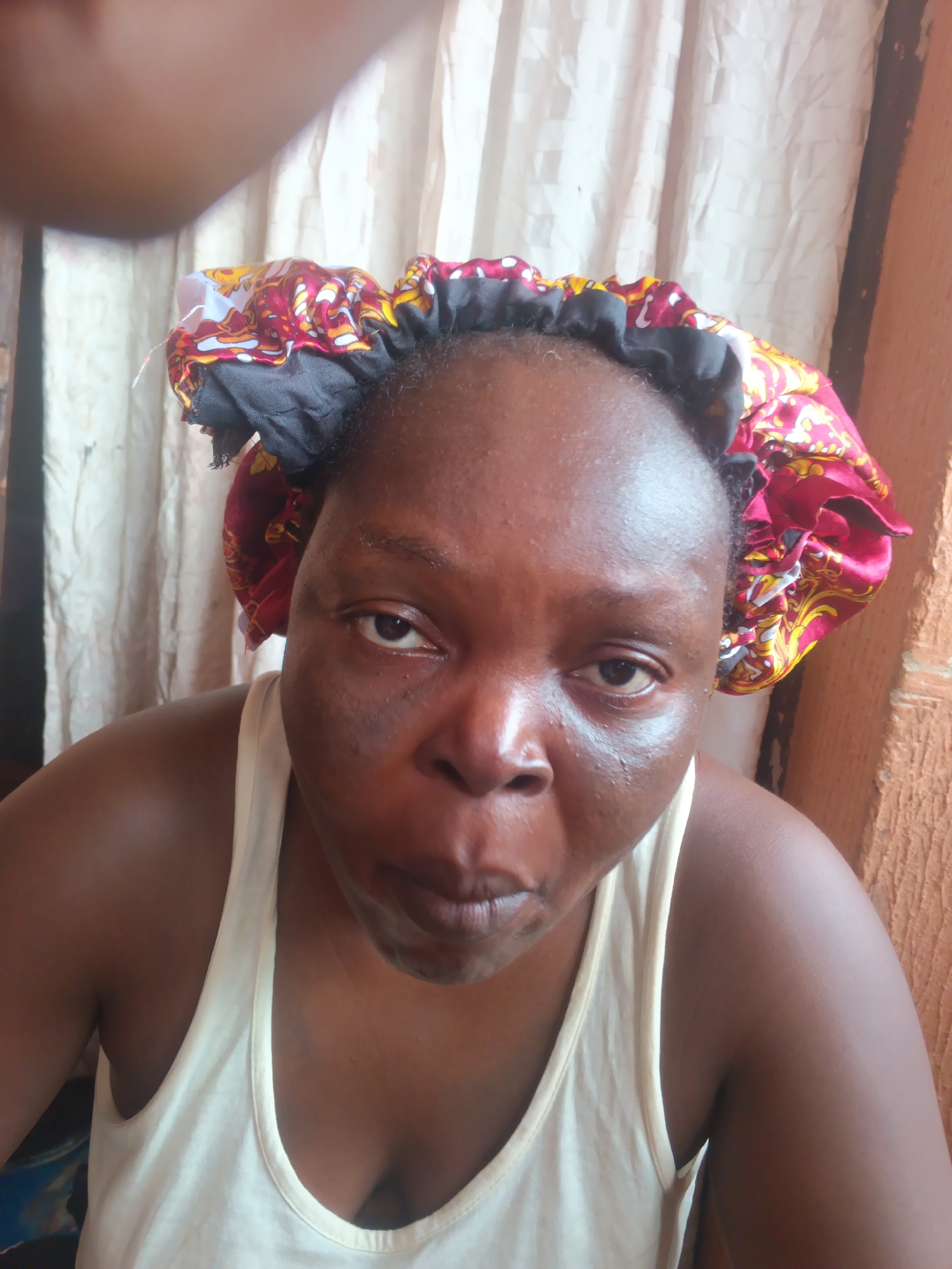 A close-up shot features a dark-skinned woman wearing a patterned headscarf and a white tank top. Her facial expression involves pursed lips and direct eye contact with the camera. The background consists of a white, slightly wrinkled curtain and what appears to be a brown wooden wall. There's insufficient detail to definitively establish the location as Onitsha, Nigeria.
