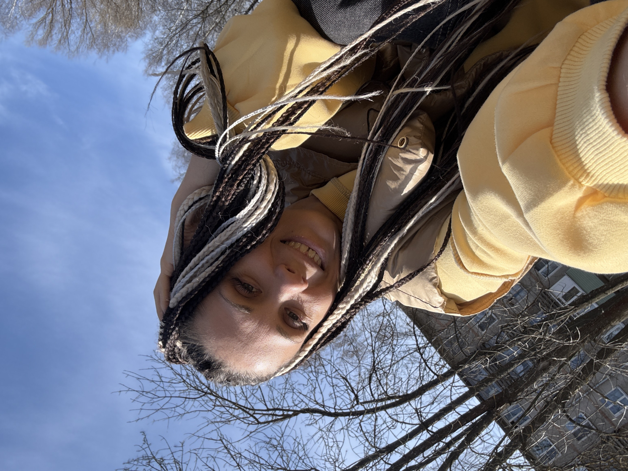 A low-angle, outdoor shot shows a woman with long, multi-toned box braids in brown, black, and white. She is smiling slightly. The braids are falling around her face and upper body. She wears a pale yellow hooded sweatshirt, over a tan jacket or vest, and a darker top. The background is filled with bare tree branches against a blue sky with scattered clouds. A multi-story building is visible in the lower right corner. The image appears to have been taken in the Rusanovo district of Russia.