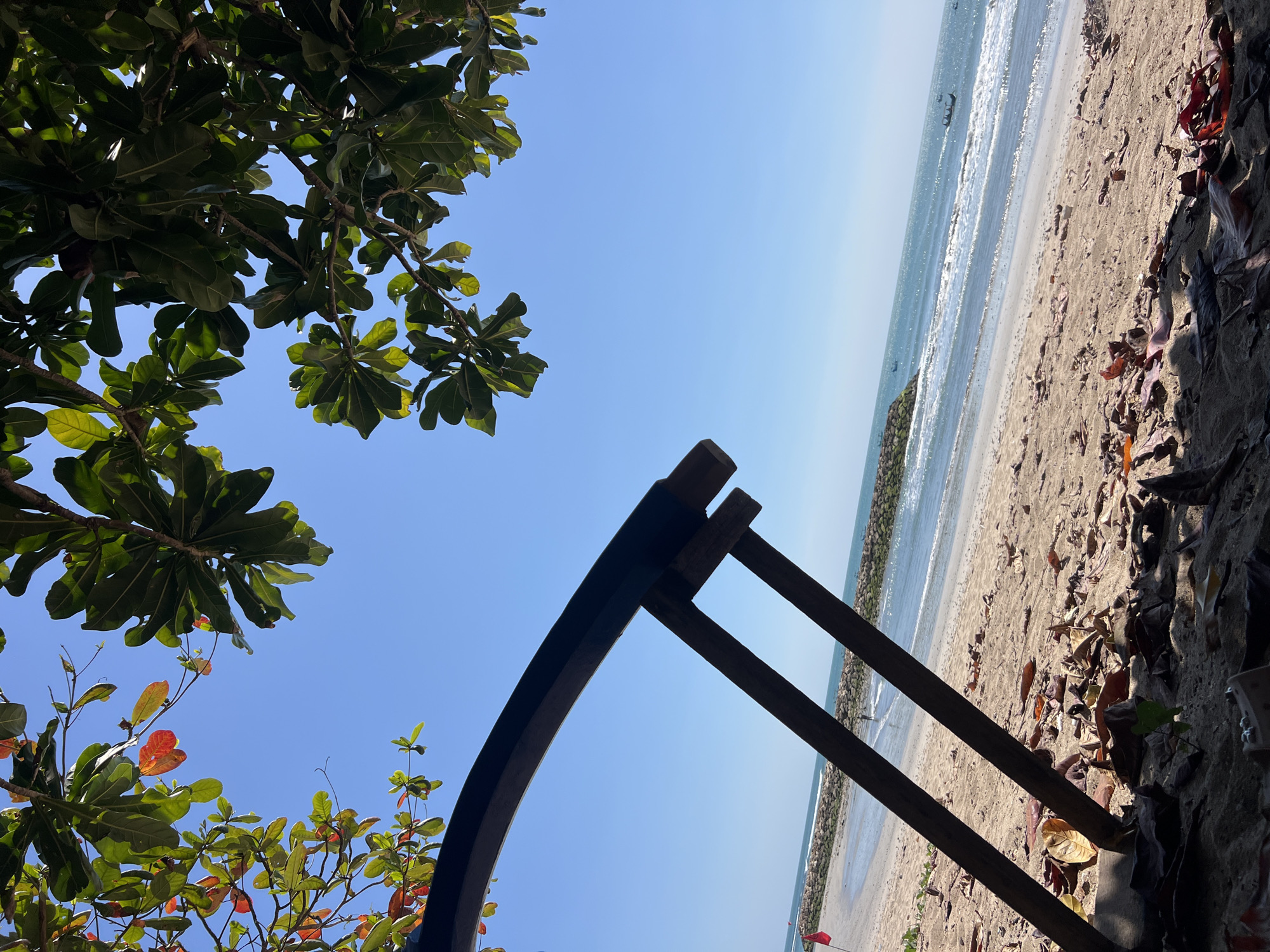 The image captures a scenic view of Kuta Beach in Bali, Indonesia, on a clear sunny day. In the foreground, a rustic wooden structure, possibly part of a beach chair or shade, is partially visible, with fallen leaves scattered on the sandy ground beneath it. The beach itself is wide and stretches towards the ocean, with gentle waves lapping the shore. A stone breakwater juts into the sea, and a few small fishing boats are dotted on the horizon. Lush green foliage from a tree hangs into the frame from the top, with some leaves displaying a touch of autumnal red, contrasting with the vibrant blue sky. The overall scene evokes a sense of tropical tranquility.