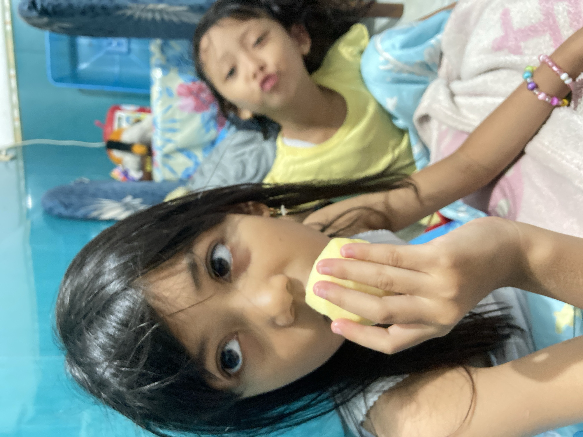 Two young girls are lying on a bed with a blue floral patterned sheet. One girl, wearing a yellow shirt, is in the background with a slightly blurred face. The other girl, in the foreground, has dark hair and is holding a yellow object, possibly food, up to her mouth. She is looking directly at the camera. The bed also has a blue and pink blanket. In the background, a blue plastic container is visible. The floor is a shiny blue. The location is Gelam, Indonesia.