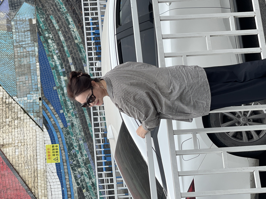 An adult female is depicted outdoors in Yuzhong District, China, positioned partially facing away from the camera, looking over her right shoulder with a slight smile. Her dark brown hair is styled in a bun, and she wears large dark sunglasses, a light-colored plaid long-sleeved shirt, dark trousers, and a gold-colored hoop earring on her visible right ear. Her right hand rests on the top rail of a white painted metal fence that extends across the mid-ground. Behind the fence, a white sedan is parked, with its rear passenger door, wheel, and trunk section partially visible. The background consists of a large wall covered in a mosaic pattern, featuring abstract designs in shades of blue, green, and white tiles, with a section of red-brown tiles on the upper left. A yellow rectangular warning sign with black text in Chinese, indicating "Warm Reminder," "Falling Objects from Height," and "Pay Attention to Safety," along with a triangular warning symbol, is affixed to the mosaic wall.