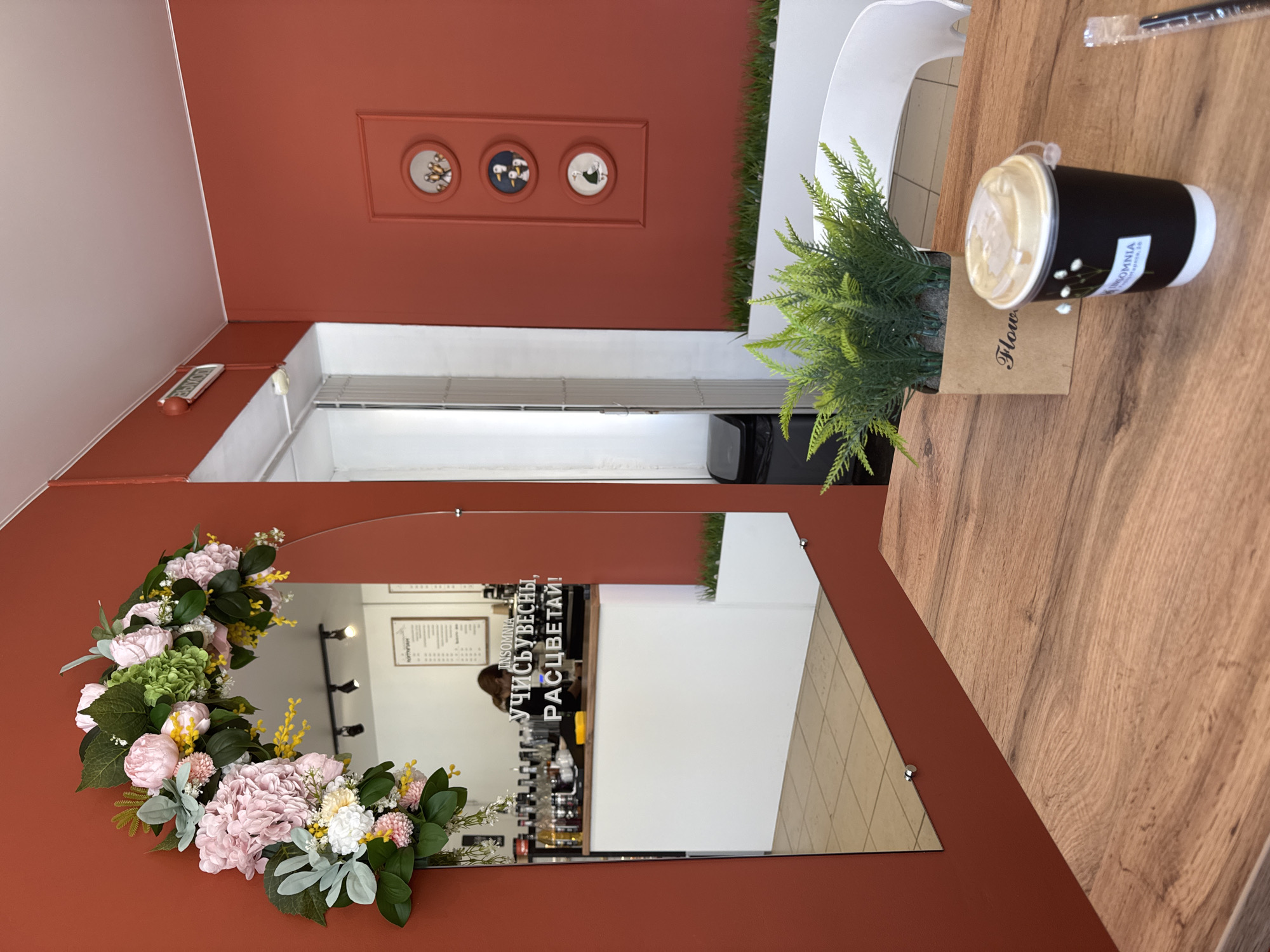 The image depicts the interior of a cafe in Chaikovsky, Russia. The perspective is from a patron's table, looking towards a mirror and a wall. The room features reddish-orange walls and a white ceiling. A large, vertically mounted rectangular mirror with rounded top corners occupies the left side of the frame, adorned with a decorative artificial floral arch featuring pink, green, and yellow flowers.
Reflected in the mirror is a coffee bar area with a white counter, a dark wooden countertop, and shelves stocked with numerous bottles. A menu board is visible on the wall behind the counter. One individual with brown hair is reflected behind the counter, facing away from the camera, consistent with a barista. Text on the mirror reads "INSOMNIA" and "УЧИСЬВЕСНЫ, РАСЦВЕТАЙ!" (Learn Spring, Bloom!). An "ВЫХОД" (Exit) sign is mounted above a doorway to the upper left, near the ceiling. A black trash bin is also visible in the reflection near the doorway. The floor in the reflection consists of light-colored tiles.
The right wall features three circular framed pictures, each containing a stylized depiction of birds, arranged vertically within a recessed rectangular frame. Below these frames, a white planter box contains artificial green grass.
In the foreground, a light-brown wooden table surface holds a small artificial potted plant in a brown paper-like container labeled "Flowers" and a black disposable coffee cup with a white base and a translucent lid. The cup is labeled "INSOMNIA" with a small floral graphic. A black pen and a clear plastic straw wrapper are partially visible to the right of the cup. A portion of a white chair's backrest is visible in the background to the right of the table.